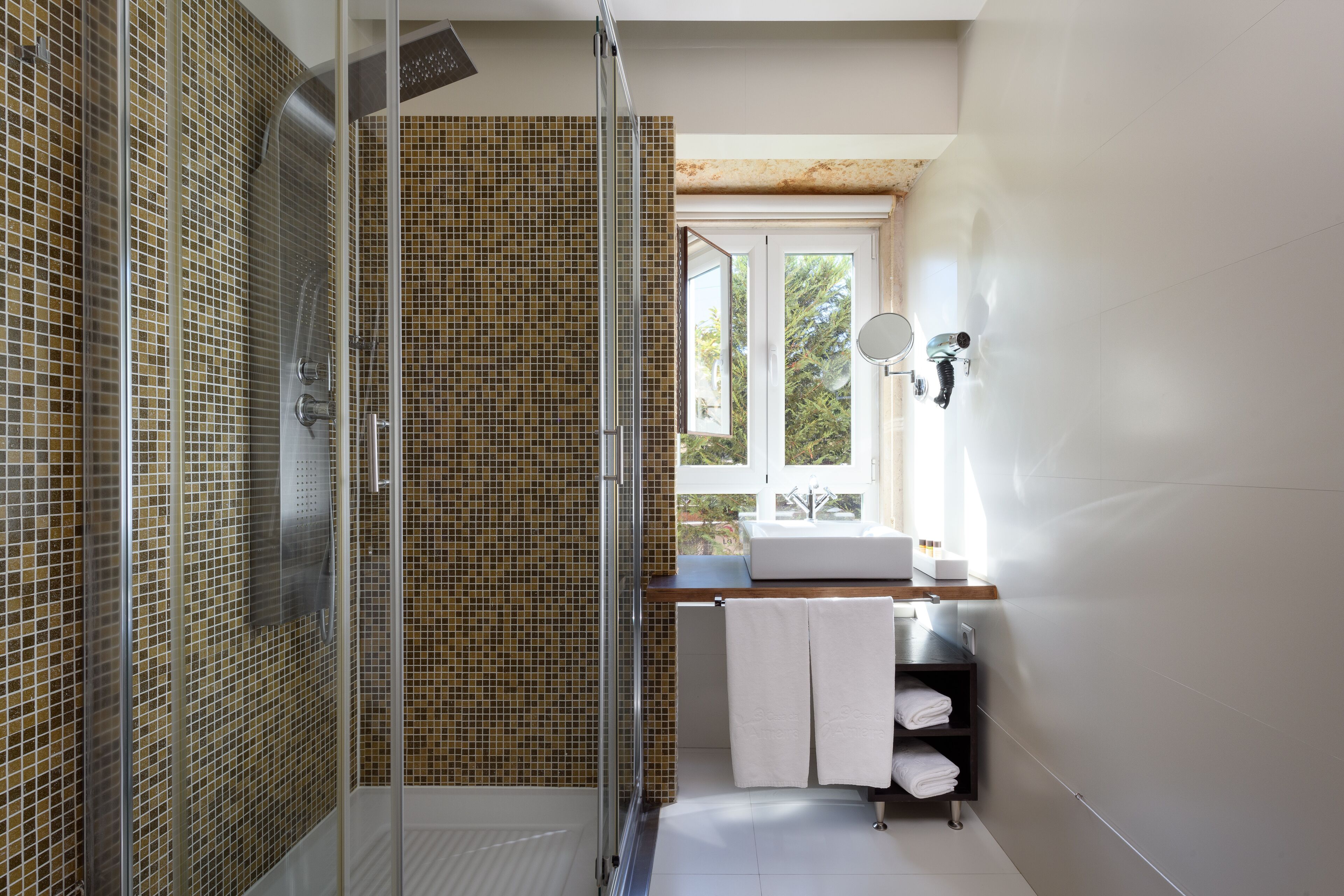 A modern bathroom with a glass shower enclosure, a tiled wall, a sink with a mirror, and a window providing natural light.