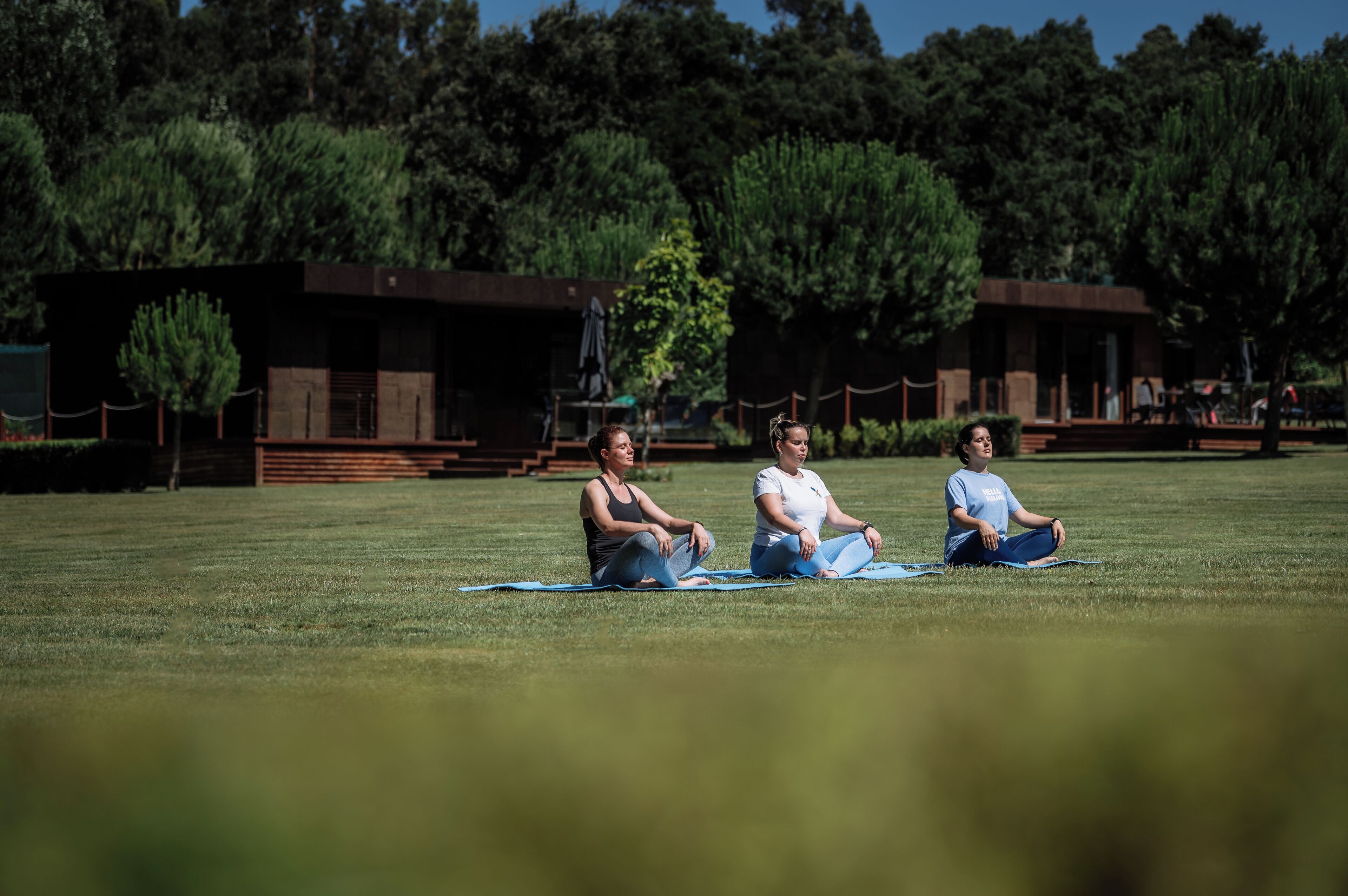 Three people sitting on mats in a grassy area with trees and buildings in the background.