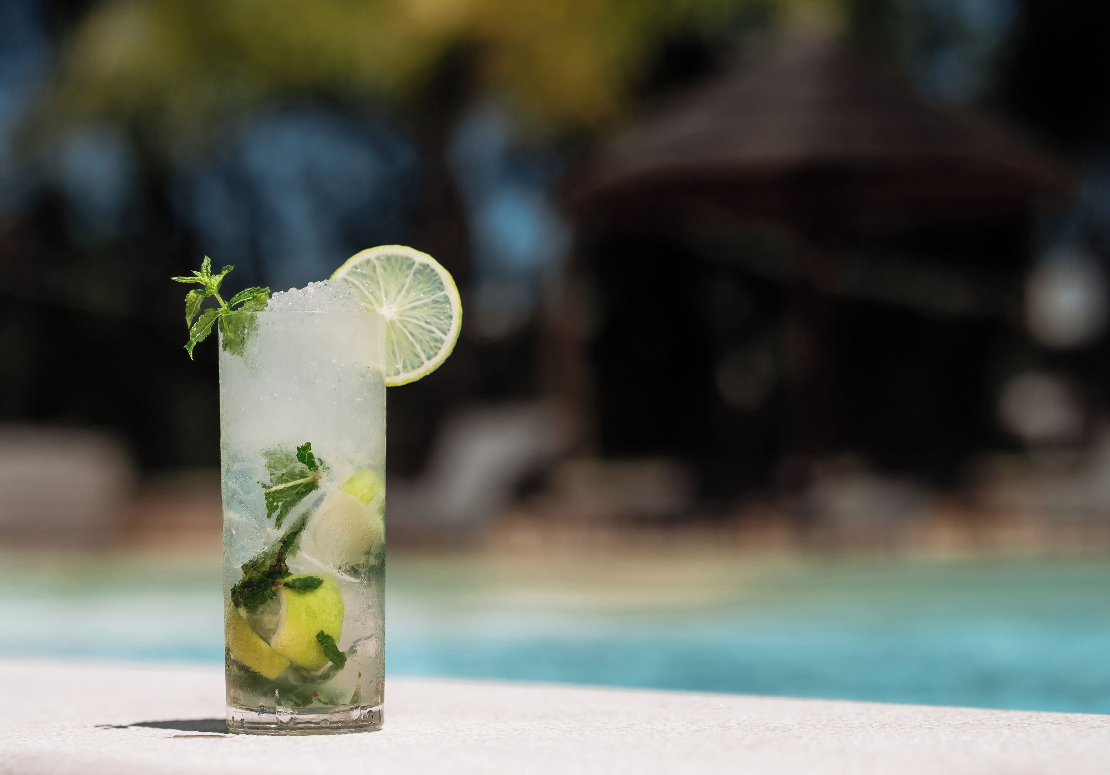 A tall glass with a lime slice and mint leaves, placed on a white surface near a pool.