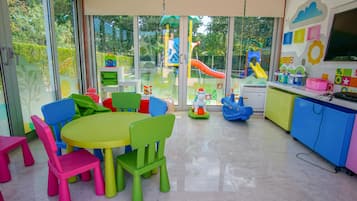 Children can play and learn at this vibrant playroom with a large table and colorful chairs, a TV, and a view of the outdoor playground.