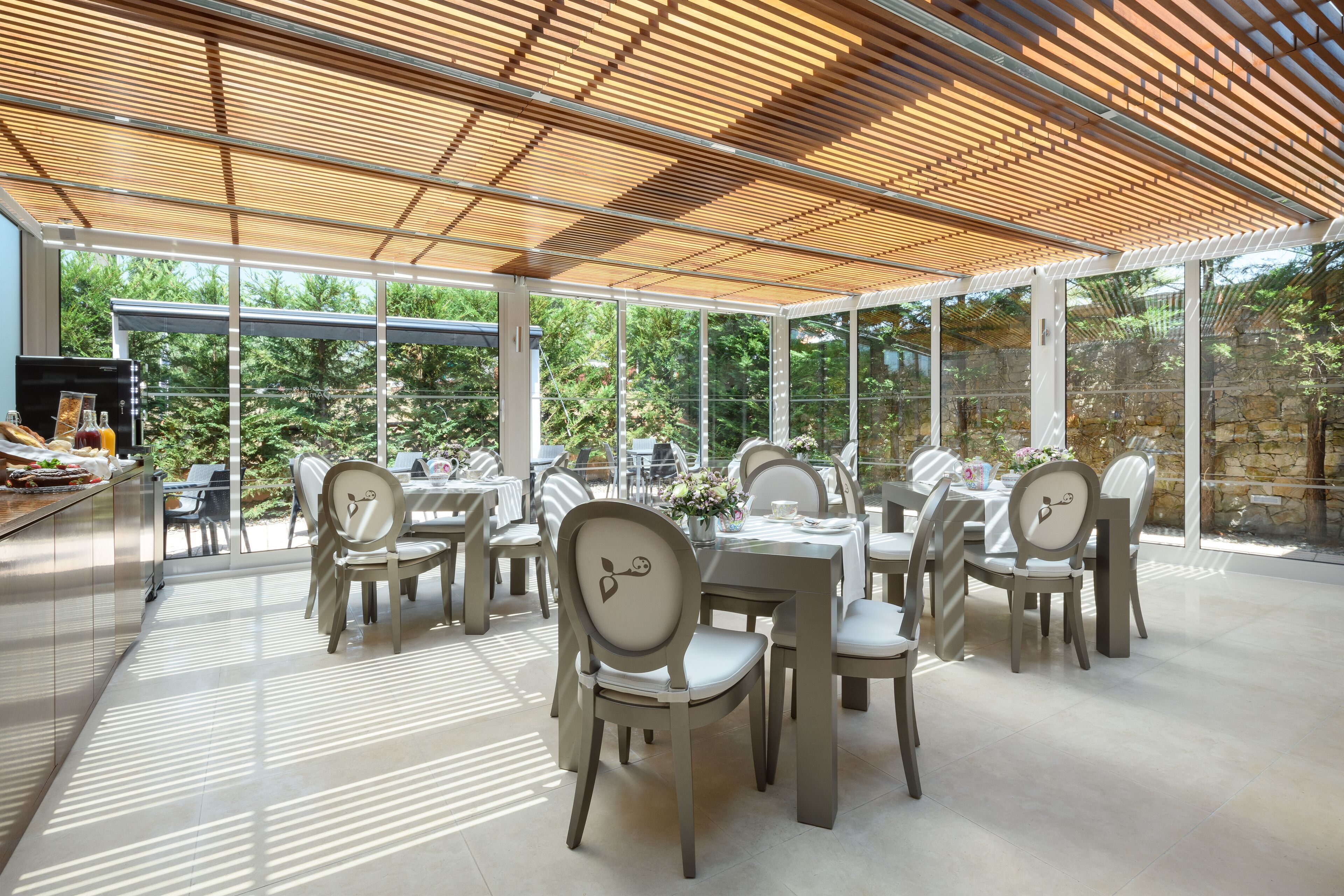 Un área de comedor al aire libre con mesas y sillas, ventanas amplias y una pérgola de madera.
