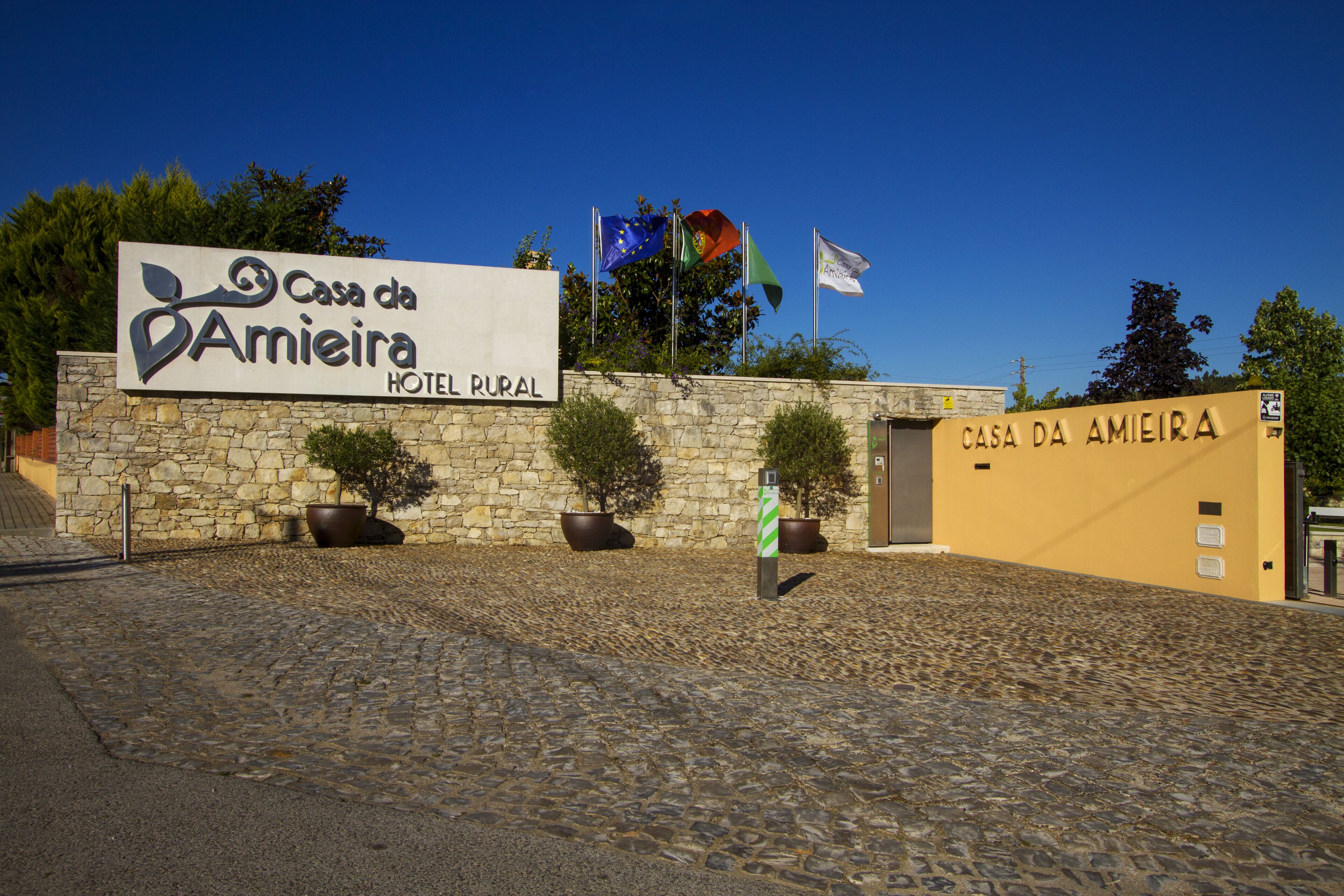 Fachada de piedra con un letrero del Hotel Rural Casa da Amieira, con plantas en macetas y banderas.