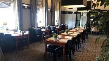 A hotel dining area with long wooden tables, black chairs, and potted plants.