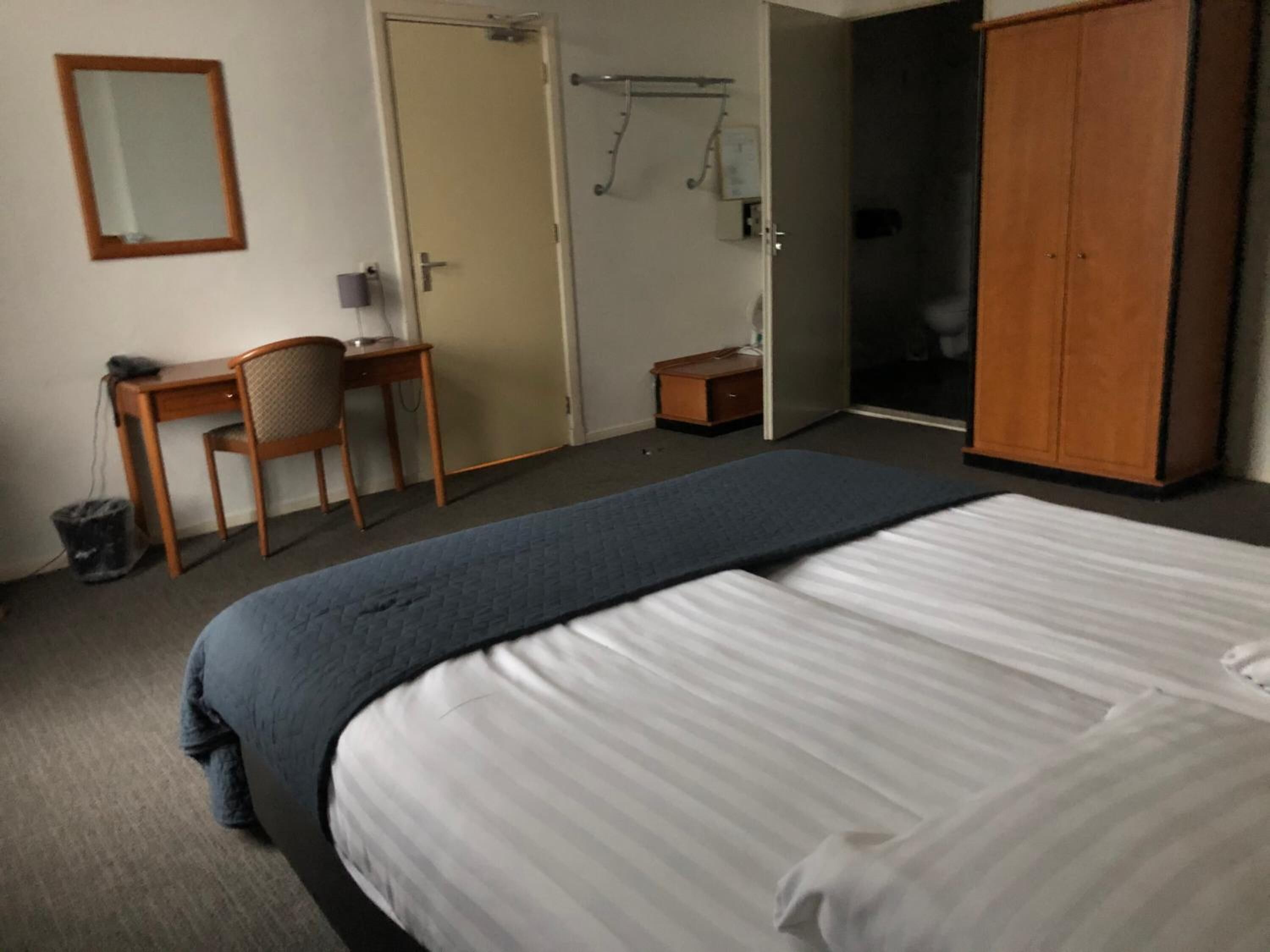 A hotel room with a bed, a desk with a chair, a wardrobe, and a mirror on the wall.