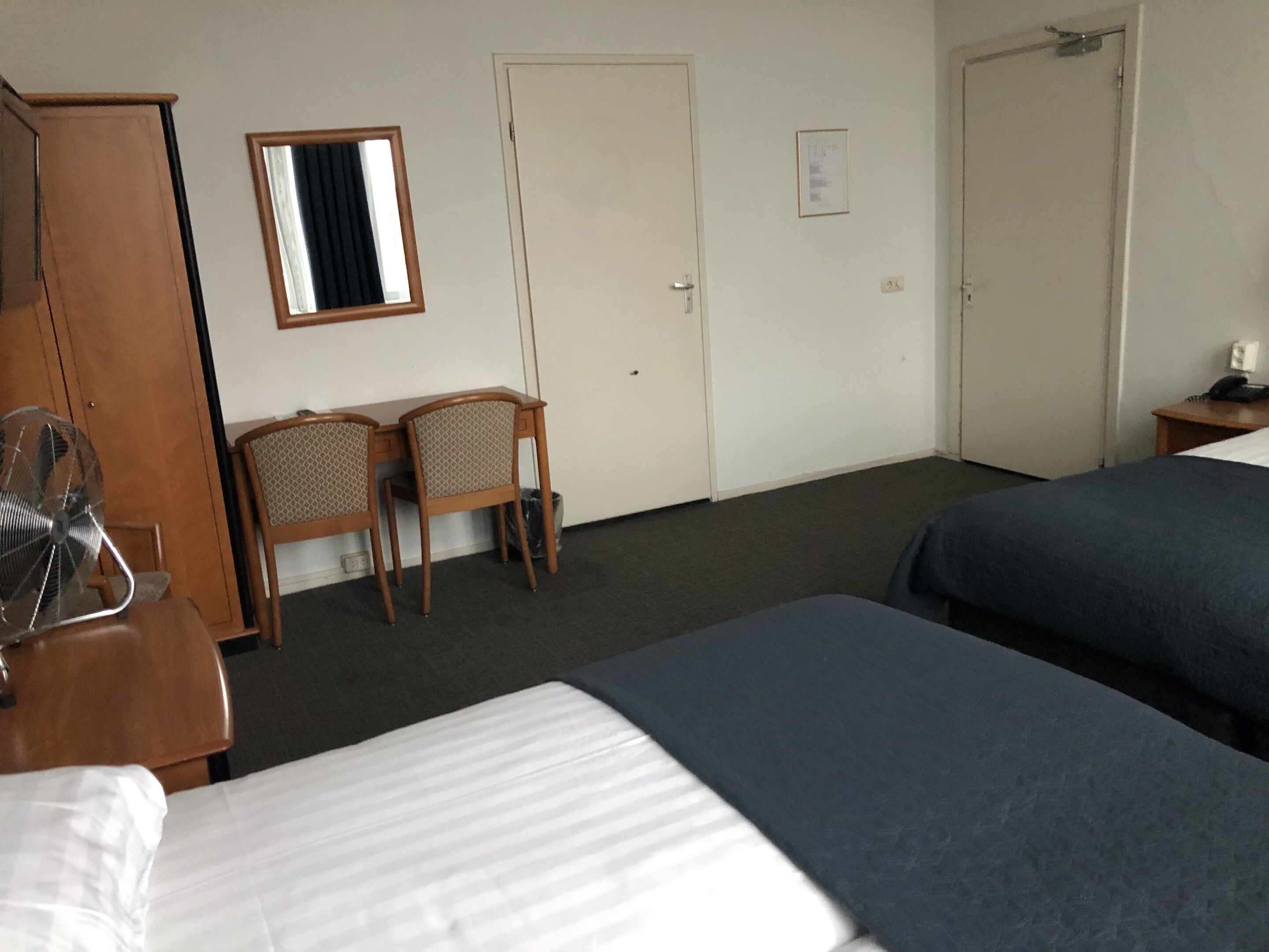 Et hotelværelse med to senge, et skrivebord med stole, et spejl og en ventilator.