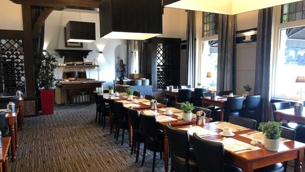 A hotel dining area with multiple tables and chairs, a kitchen in the background, and large windows allowing natural light. - Hotel Wilhelmina (Venlo)