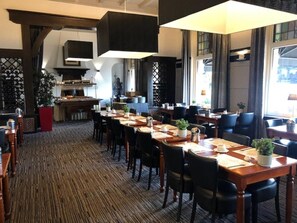 Een hotelrestaurant met meerdere tafels en stoelen, een keuken op de achtergrond en grote ramen voor natuurlijk licht.