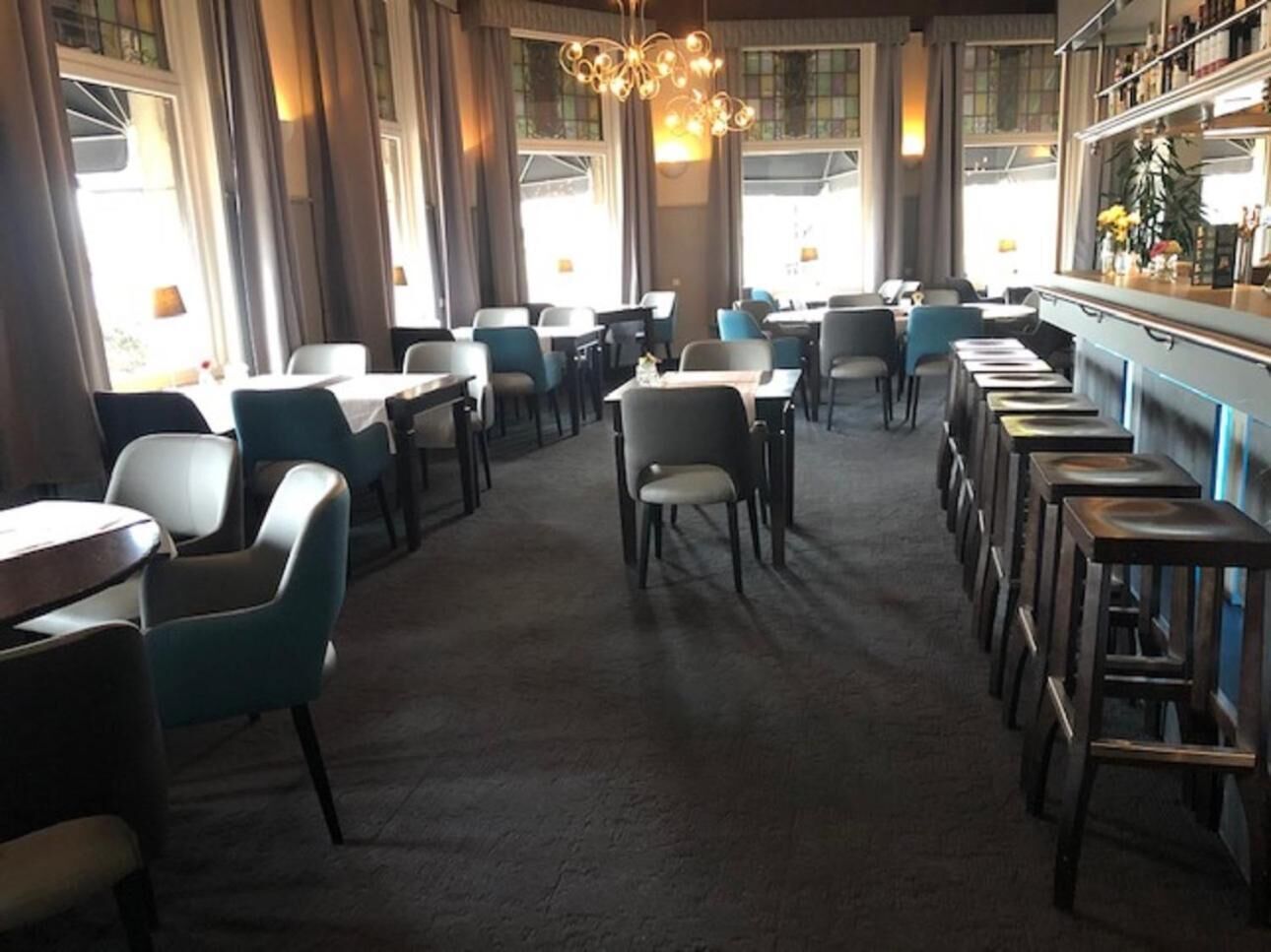 Un restaurante con zona de bar, varias mesas y sillas, y amplios ventanales que dejan entrar luz natural.