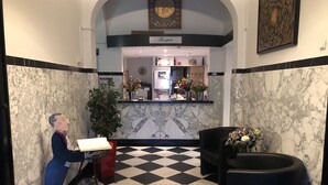 A lobby with a checkered floor, marble walls, a reception desk, a statue, and a reception area with flowers and a clock. - Hotel Wilhelmina (Venlo)