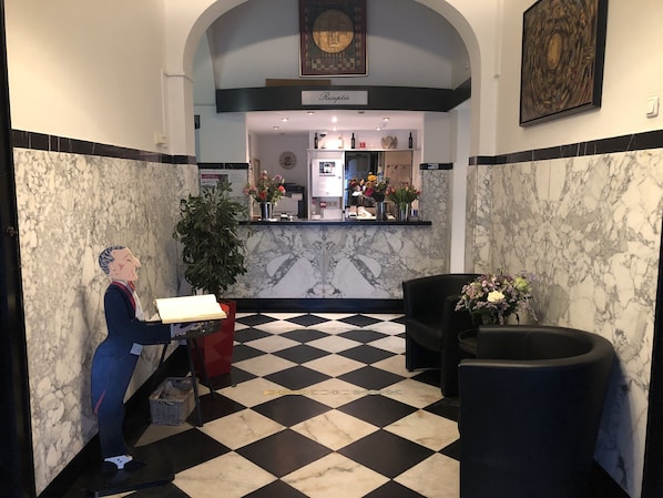 A lobby with a checkered floor, marble walls, a reception desk, a statue, and a reception area with flowers and a clock. - Hotel Wilhelmina (Venlo)