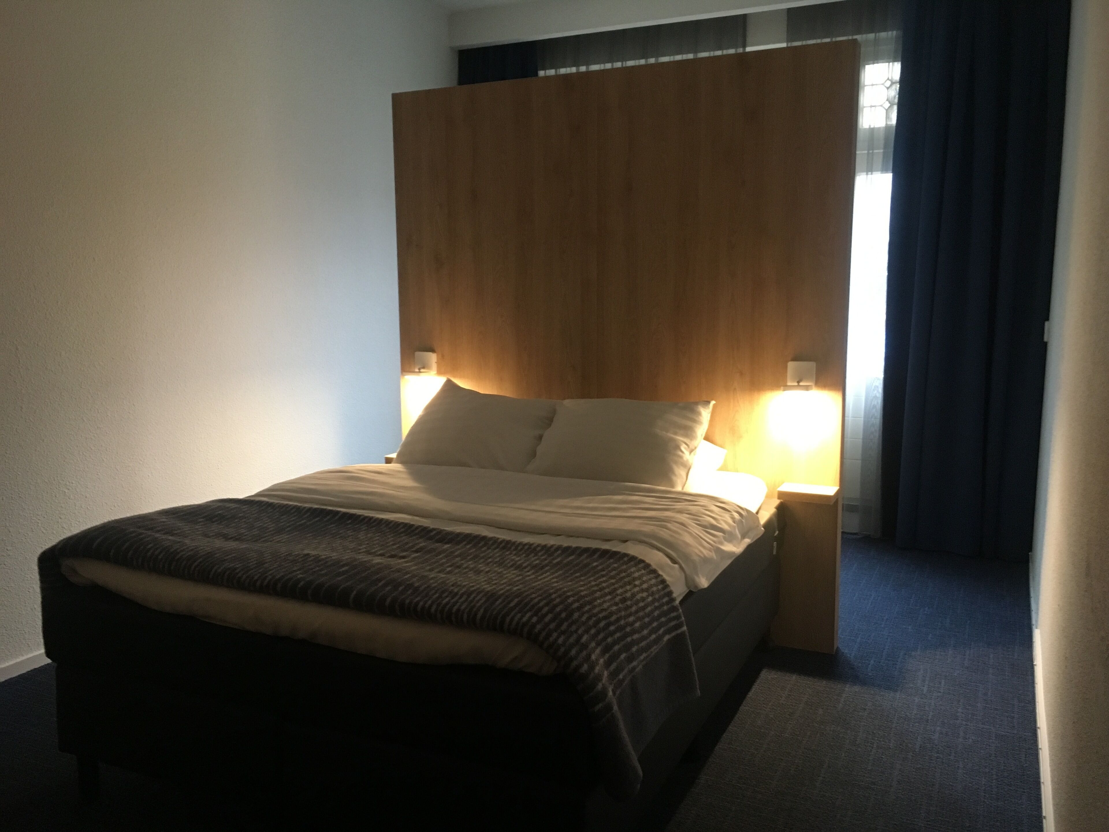 A neatly made bed with a headboard, two bedside lamps, and a window with curtains.