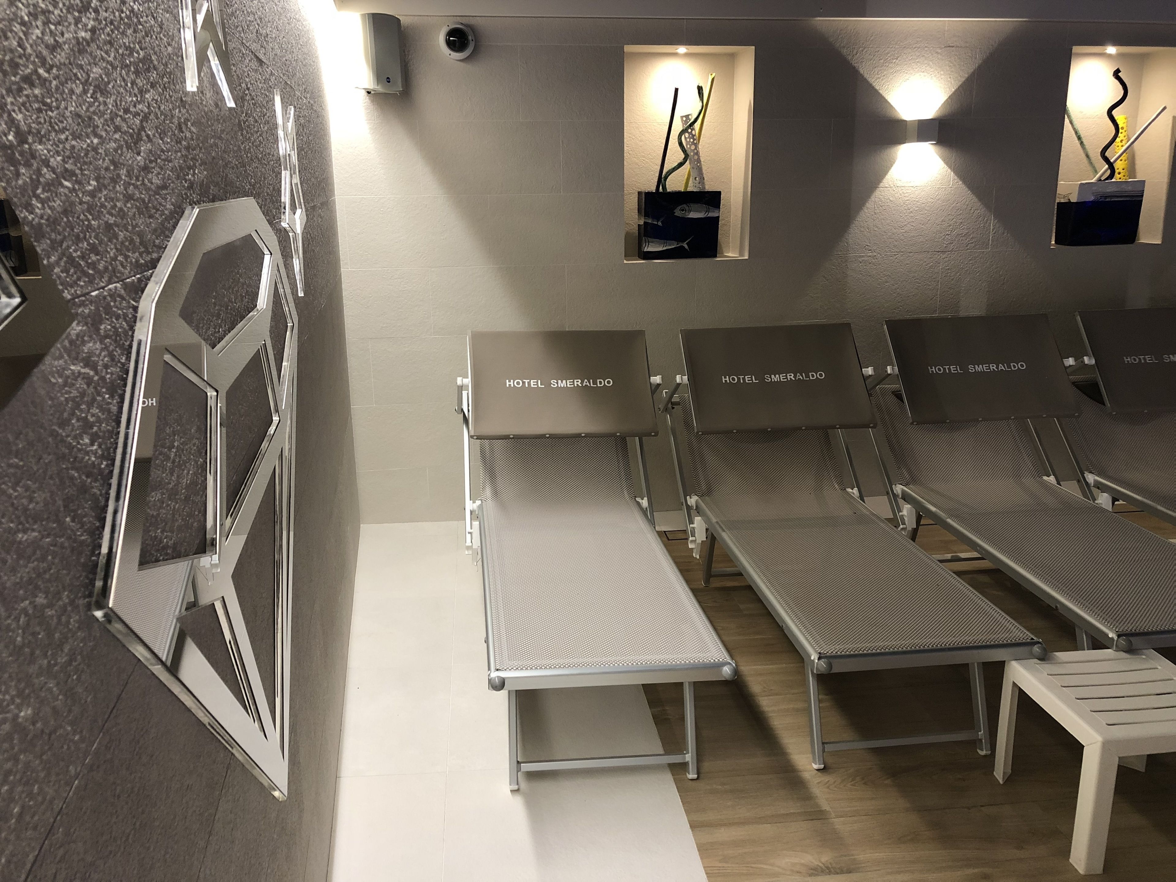 A hotel room with a modern design, featuring a seating area with lounge chairs and a small table, a wall-mounted security camera, and a framed picture of a building.