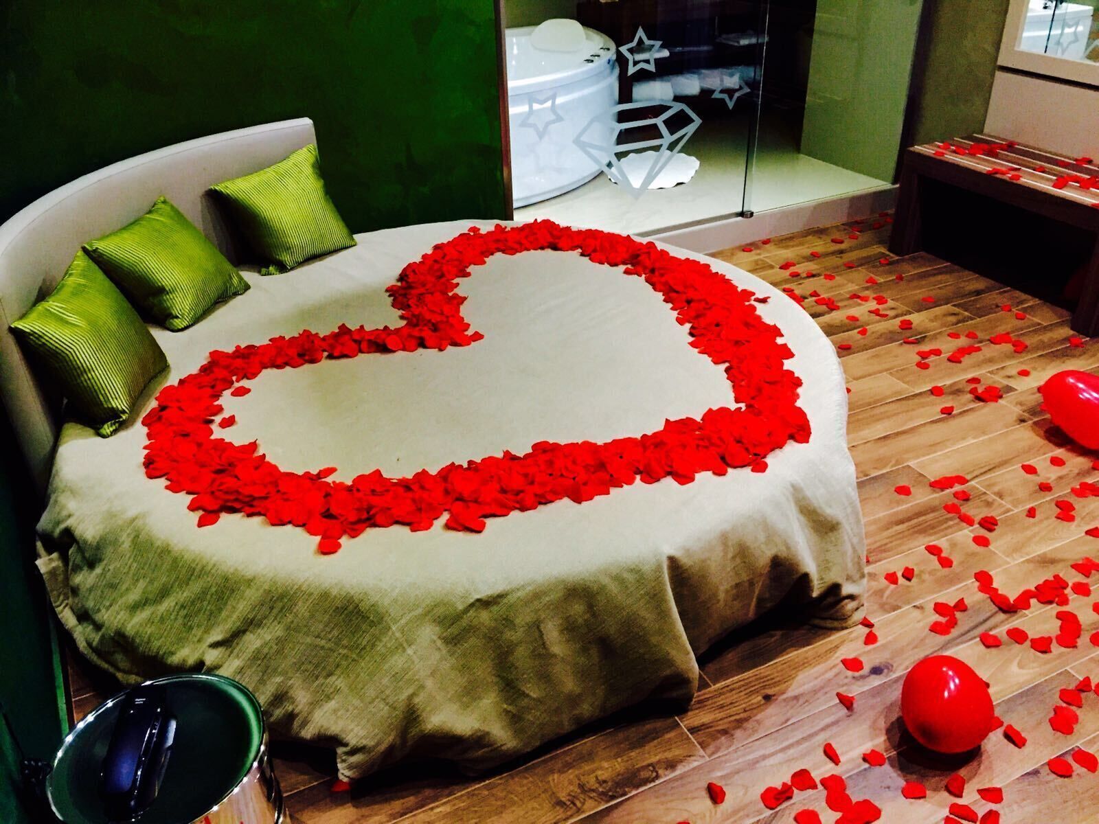 Un letto con un'elegante composizione di petali di rosa a forma di cuore sul copripiumino, cuscini verdi e un pavimento in legno spolverato di petali di rosa e palloncini.