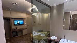 A modern bedroom with a glass partition, a television, a bed, a wooden floor, and a large bathtub. - Hotel Smeraldo (Qualiano)