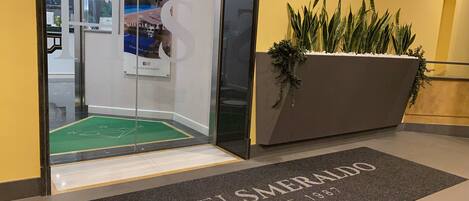 A hotel entrance with a glass door, a green mat with a logo, and a planter with green plants.
