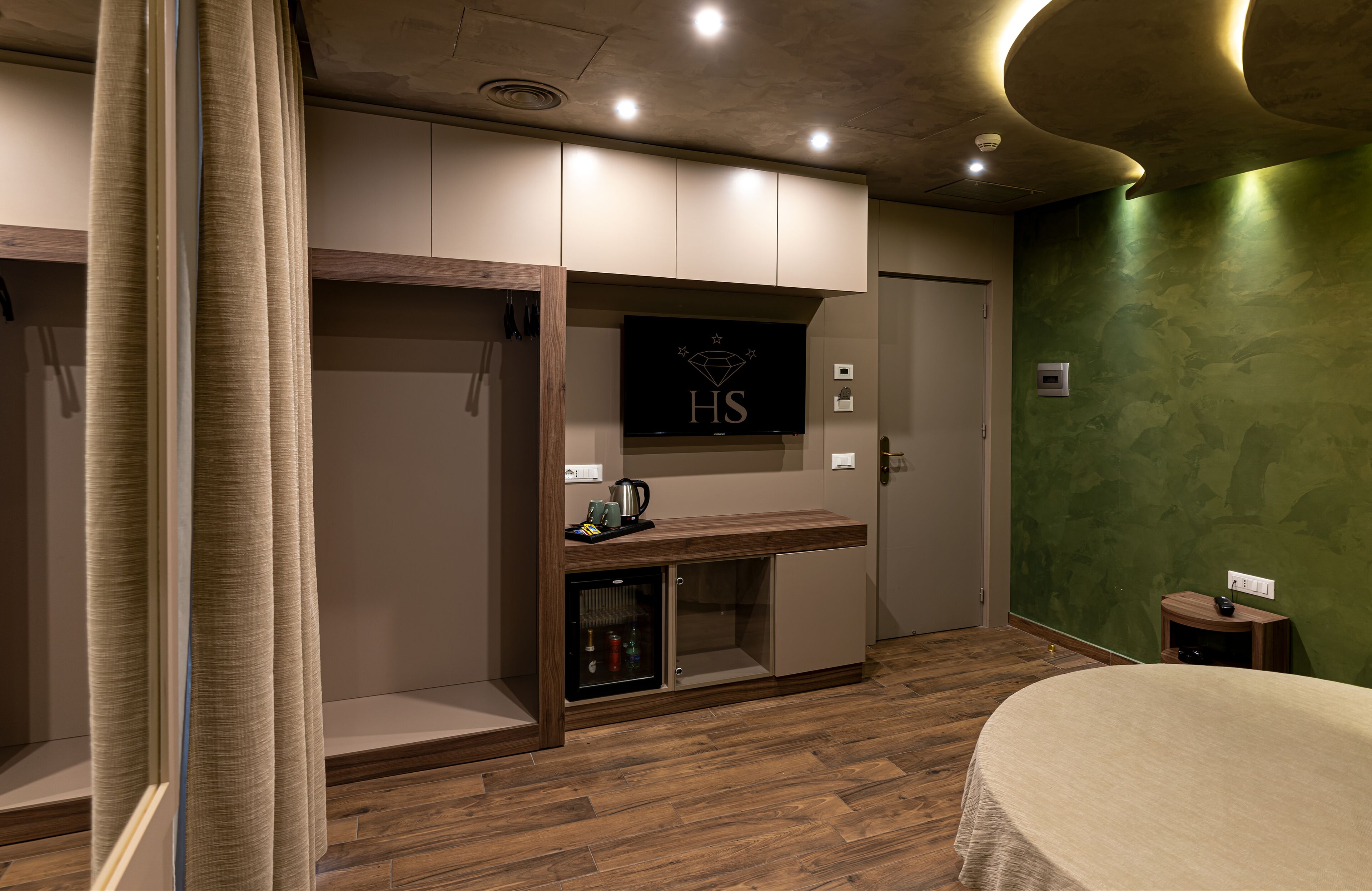 A hotel room with a wooden floor, a bed, a TV, a minibar, and a green accent wall.