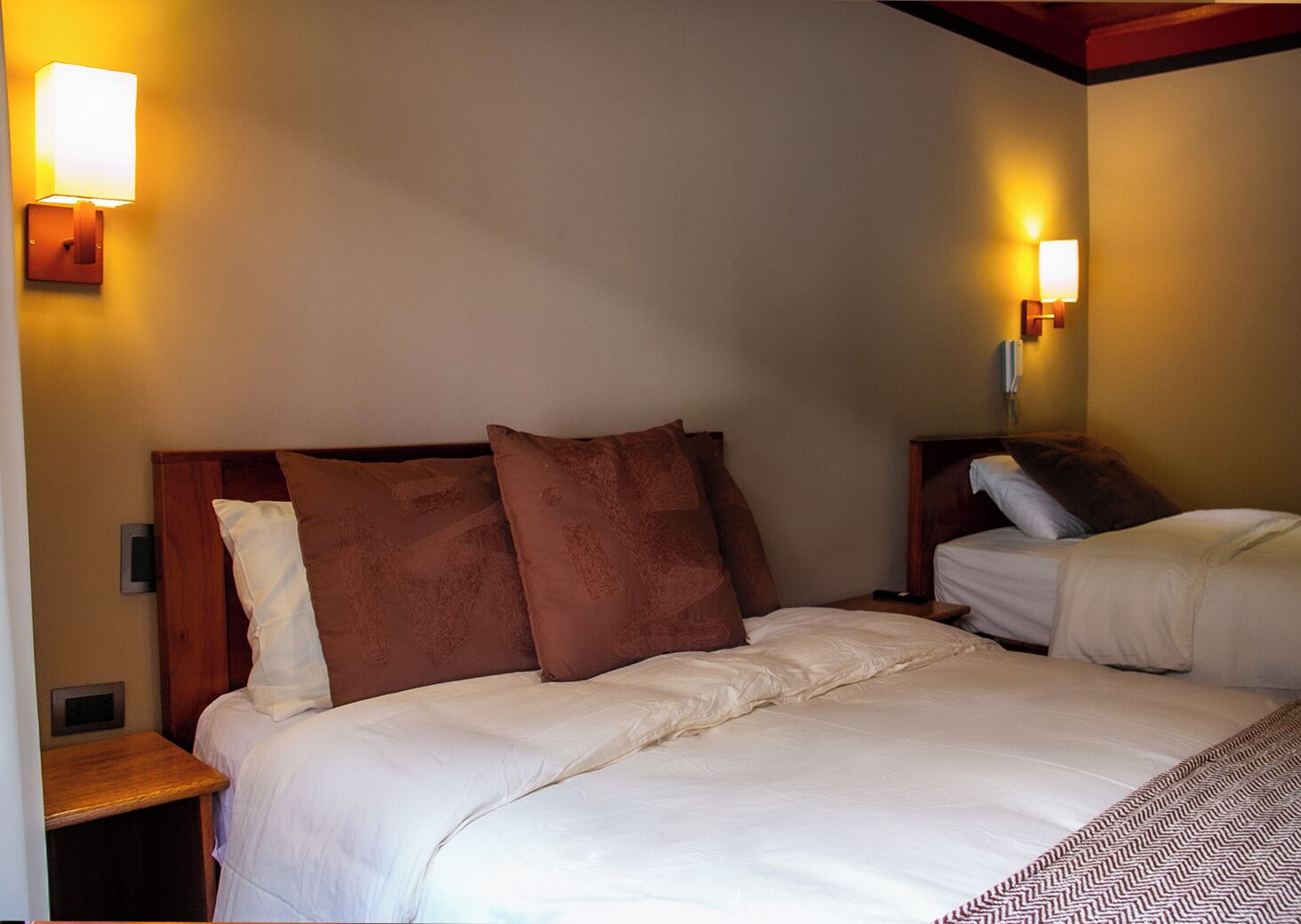Una habitación de hotel con dos camas, cada una con ropa blanca y almohadas marrones. Hay lámparas de pared a cada lado de las camas.