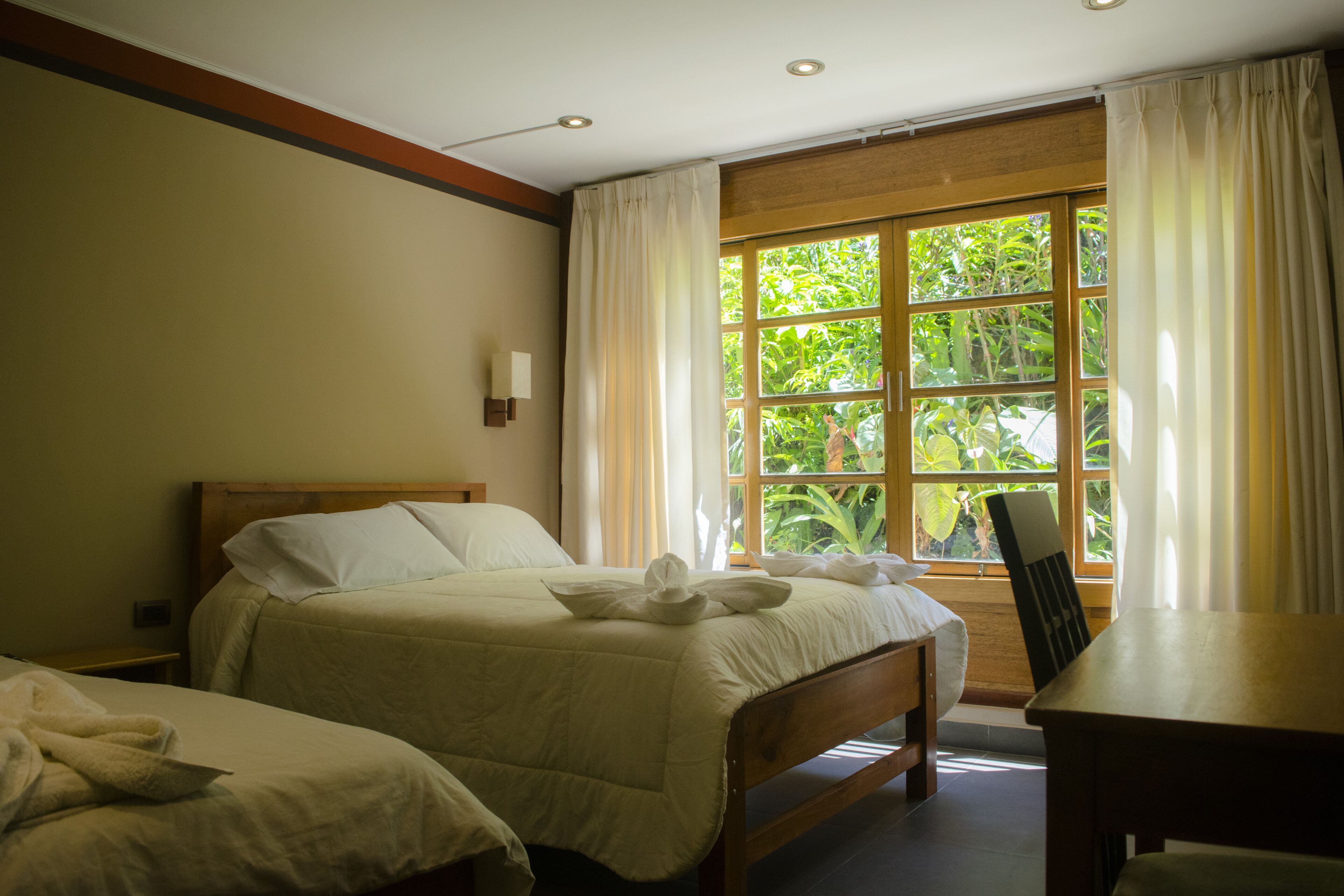 A hotel room with a large bed, a smaller bed, a wooden table, and a window with curtains.