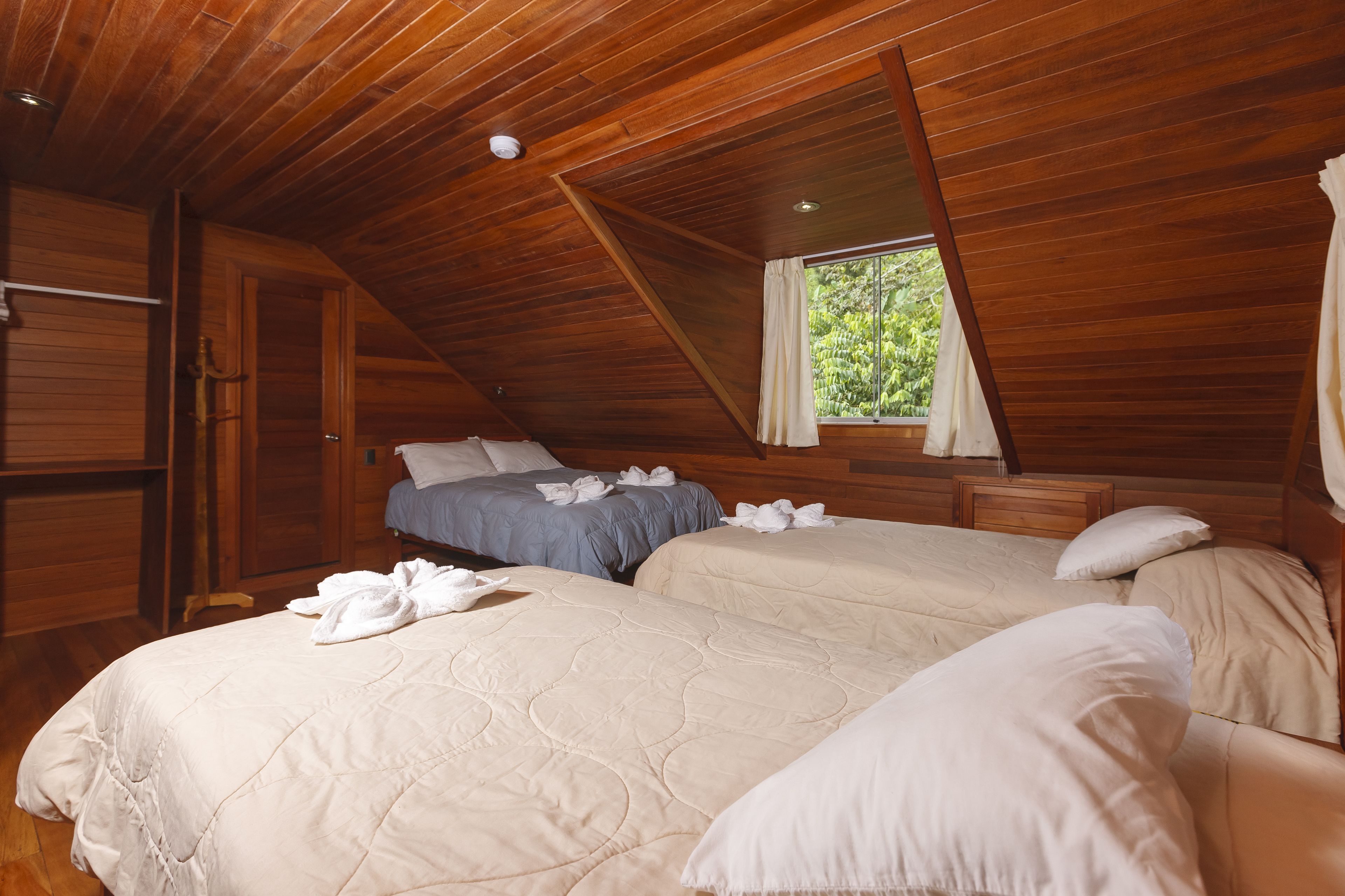 Una habitación con vigas de madera en el techo, dos camas con ropa de cama color beige, una ventana con cortinas y un armario de madera.