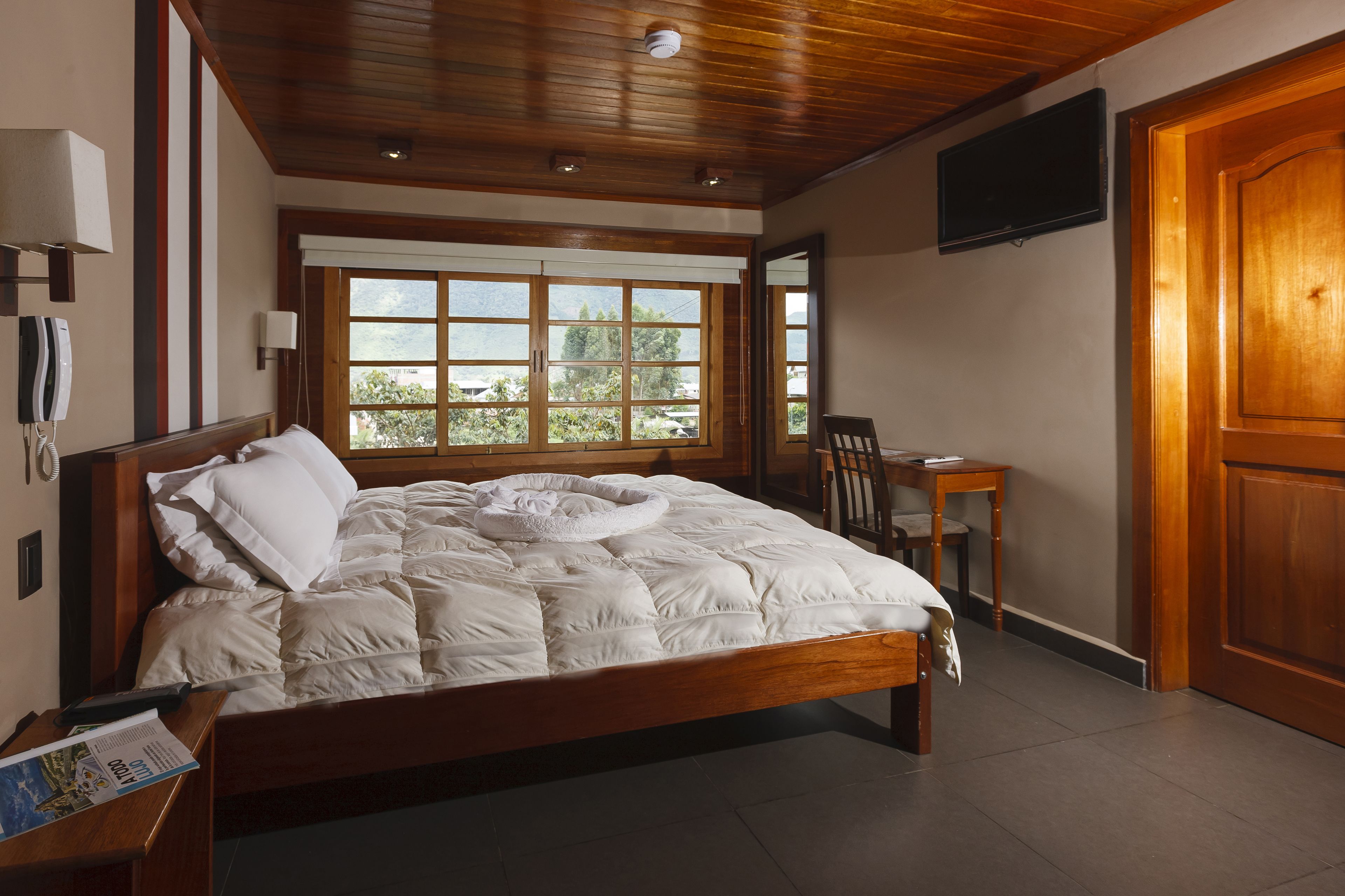 A neatly made bed with a white comforter and pillows, a wooden headboard, a bedside table with a lamp, a chair, a small table, and a TV mounted on the wall.
