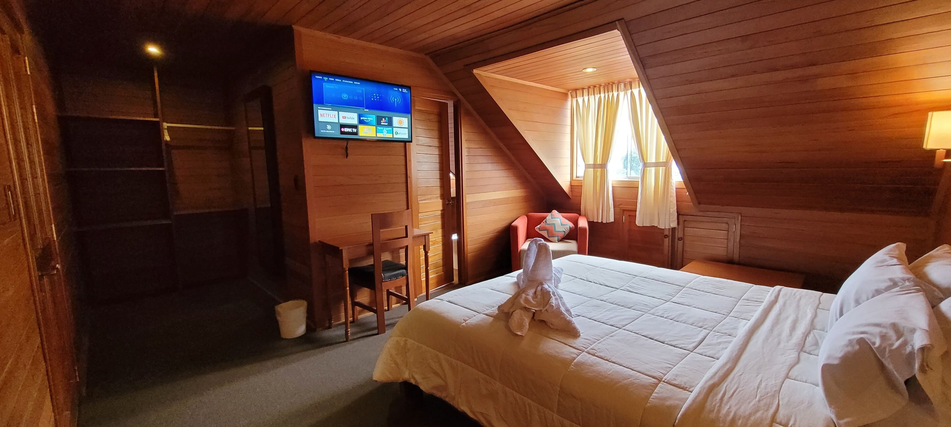 A cozy attic bedroom with a wooden sloped ceiling, a flat-screen TV mounted on the wall, a bed with white linens, and a chair with a pillow.