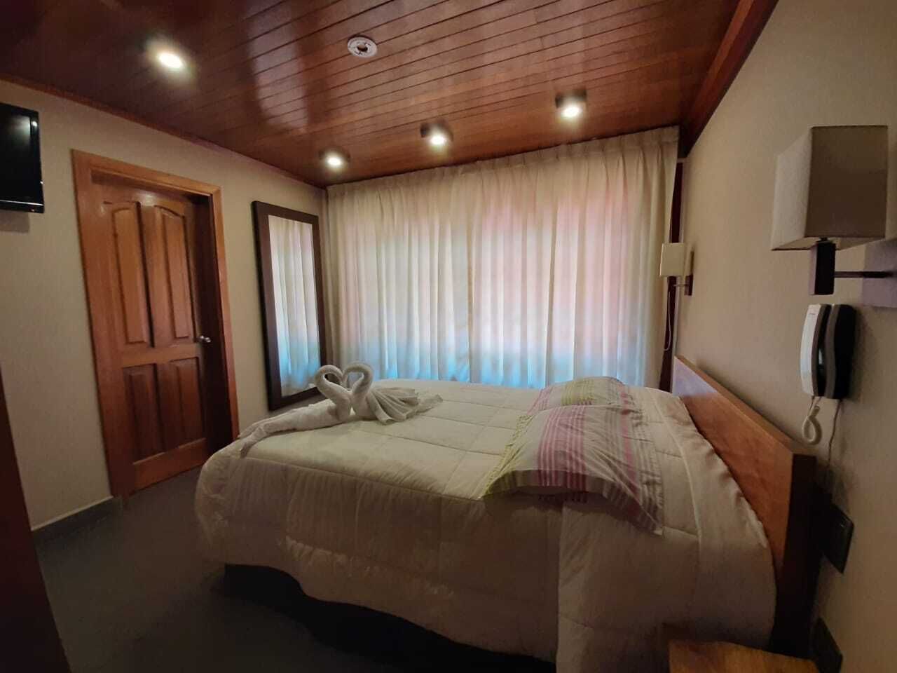 A hotel room with a wooden ceiling, a bed with a white comforter, a telephone on the wall, and a window with curtains.
