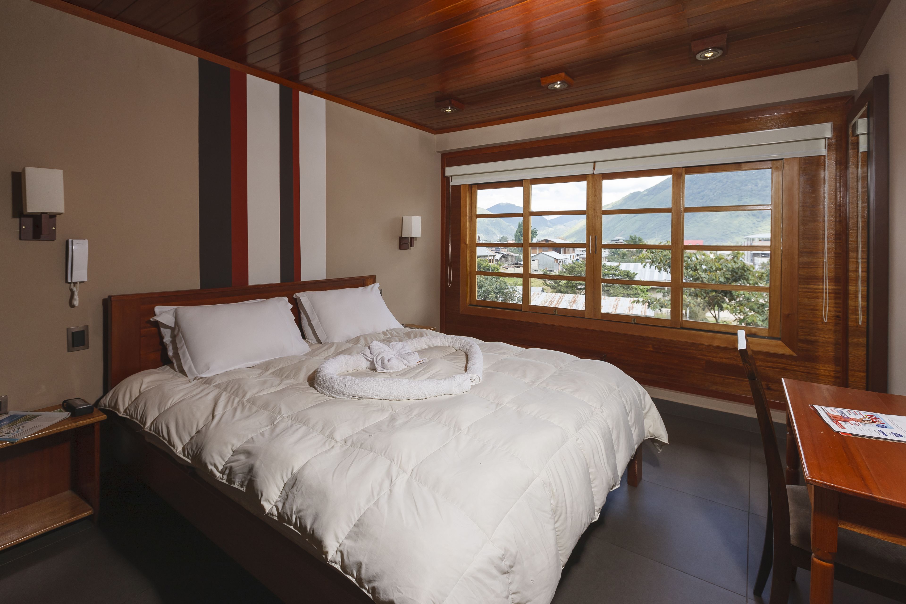 A hotel room with a large bed, wooden ceiling, a window with a view of mountains, and a wooden desk with a chair.