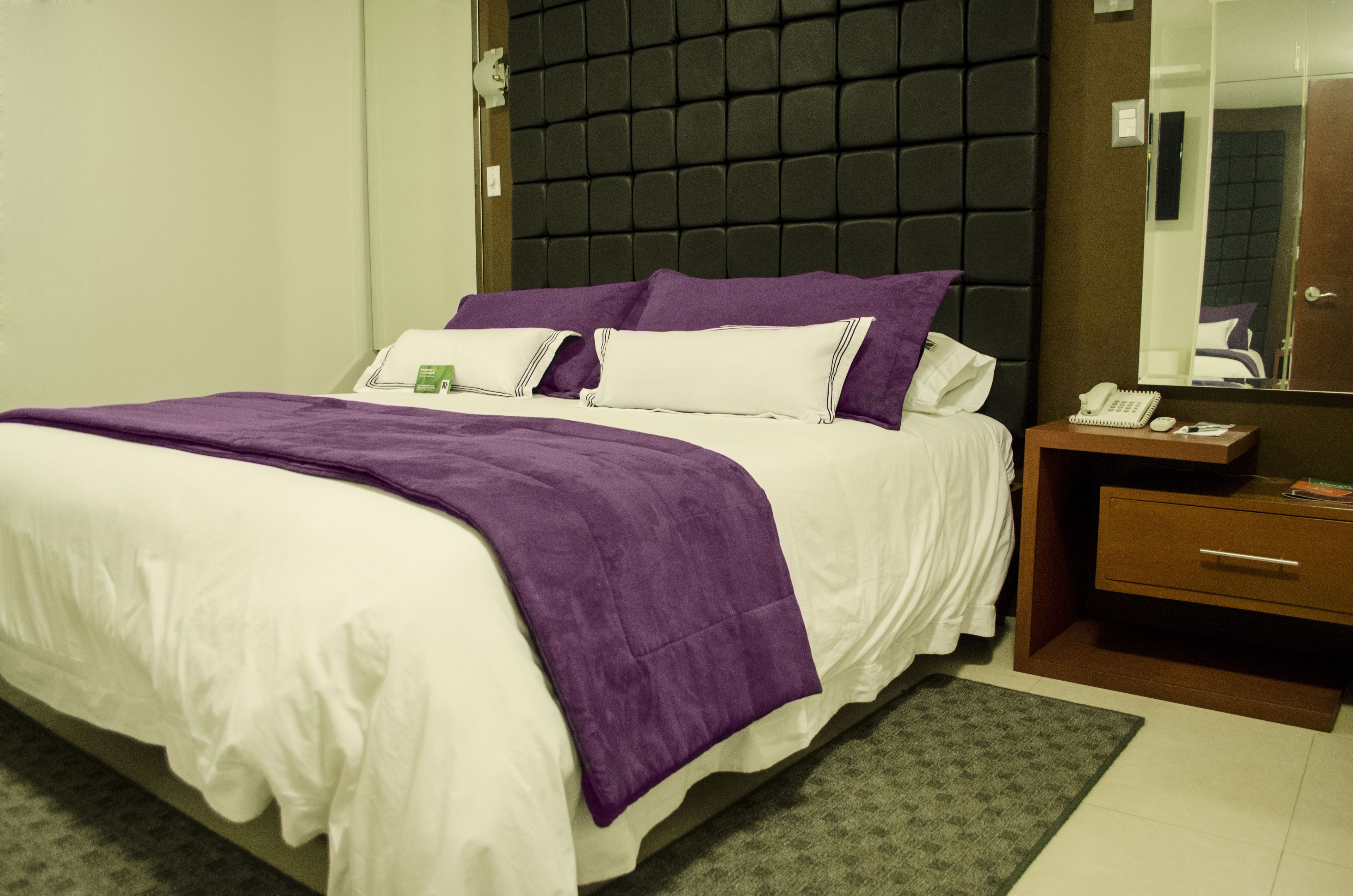 Una cama bien hecha con ropa de cama blanca y morada, un cabecero oscuro, una mesita de noche con un teléfono y una lámpara, y un espejo en la pared.
