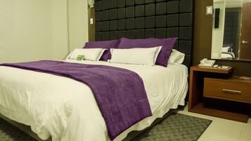 Una cama bien hecha con ropa de cama blanca y morada, un cabecero oscuro, una mesita de noche con un teléfono y una lámpara, y un espejo en la pared.