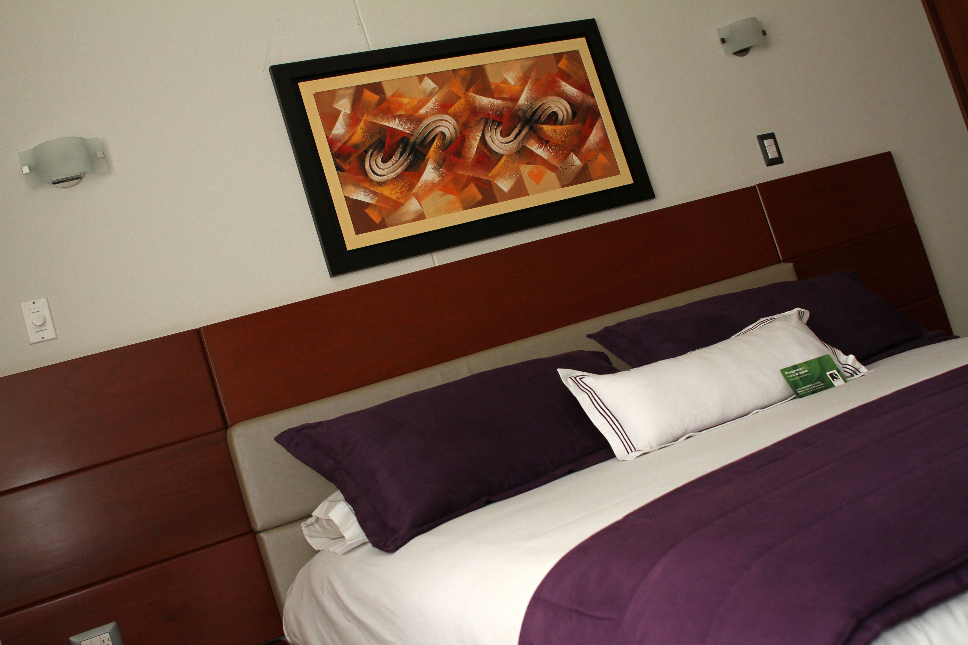 Habitación de hotel con una cama, ropa de cama púrpura y blanca, un cuadro abstracto enmarcado en la pared y un cabecero de madera.