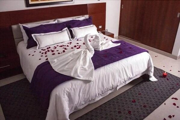 A neatly made bed with a purple blanket and white linens, a wooden headboard, and a red candle on the floor beside the bed. - Sunec Hotel (Chiclayo)