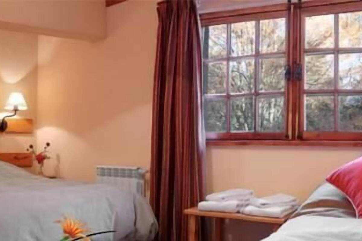 Habitación de hotel con dos camas, una mesita de noche, una lámpara y un ventanal con cortinas.