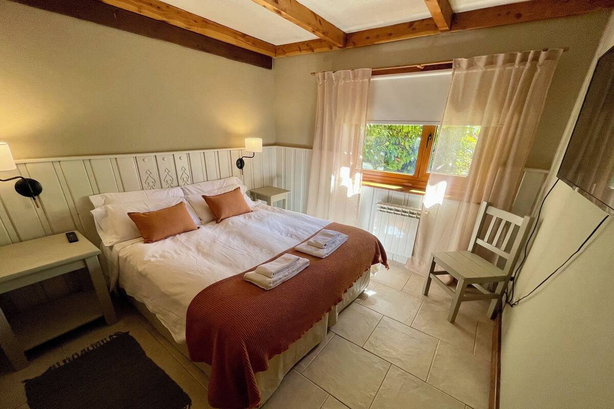 Un dormitorio con cama, mesitas de noche, una silla, ventana con cortinas y radiador.