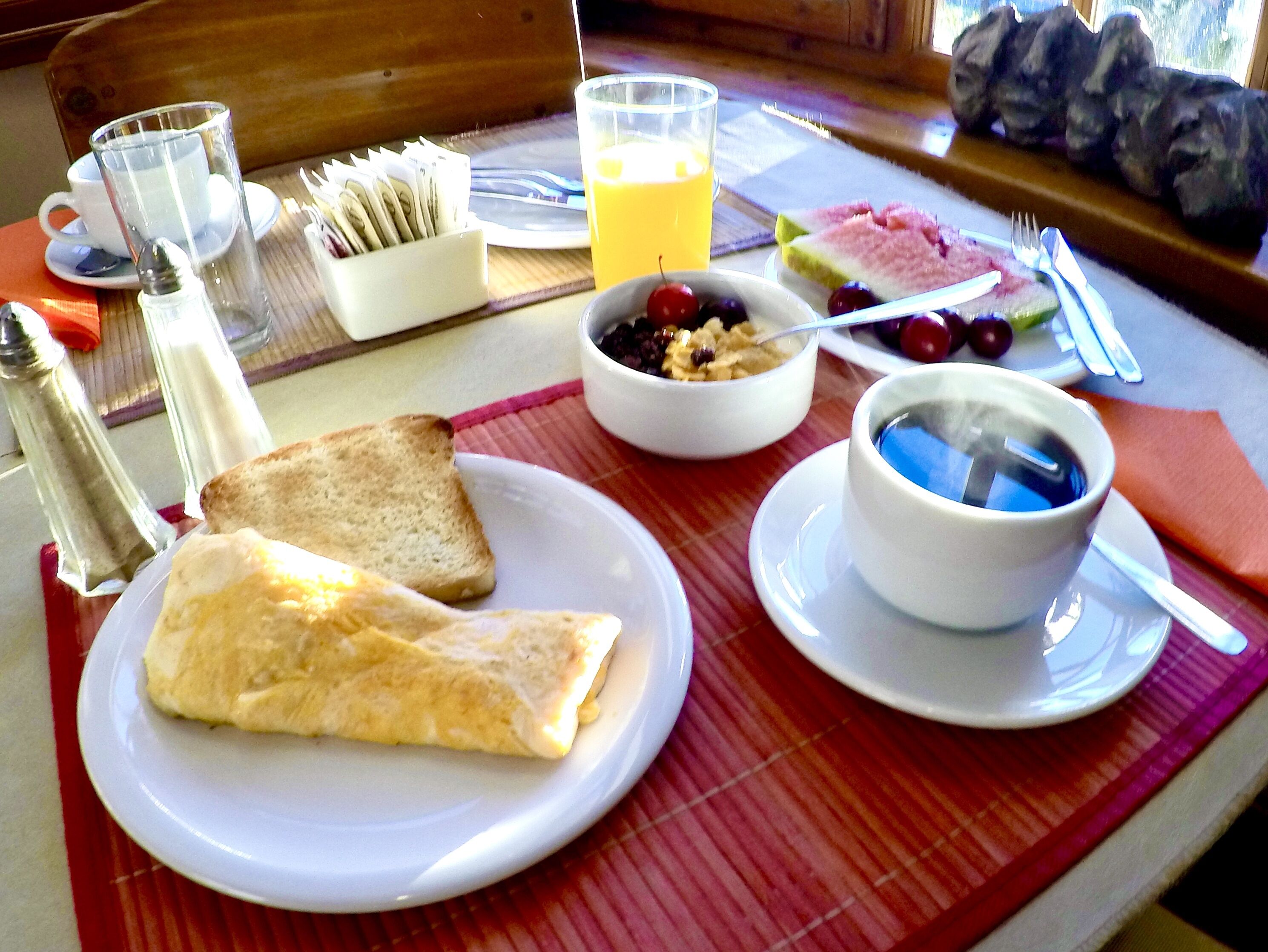 Una escena de desayuno con tostadas, un tazón de fruta, un vaso de jugo de naranja y una taza de café sobre una mesa.