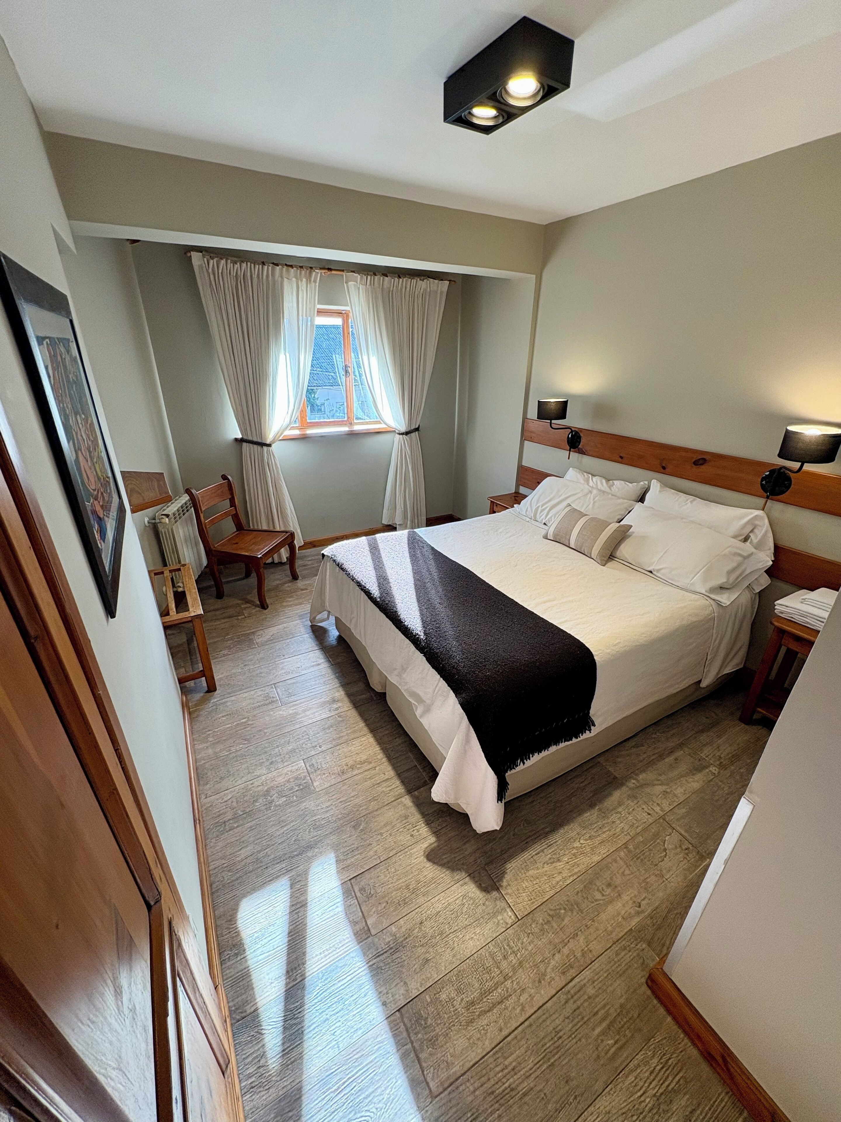 A bedroom with a large bed, a window with curtains, a chair, and a small table.