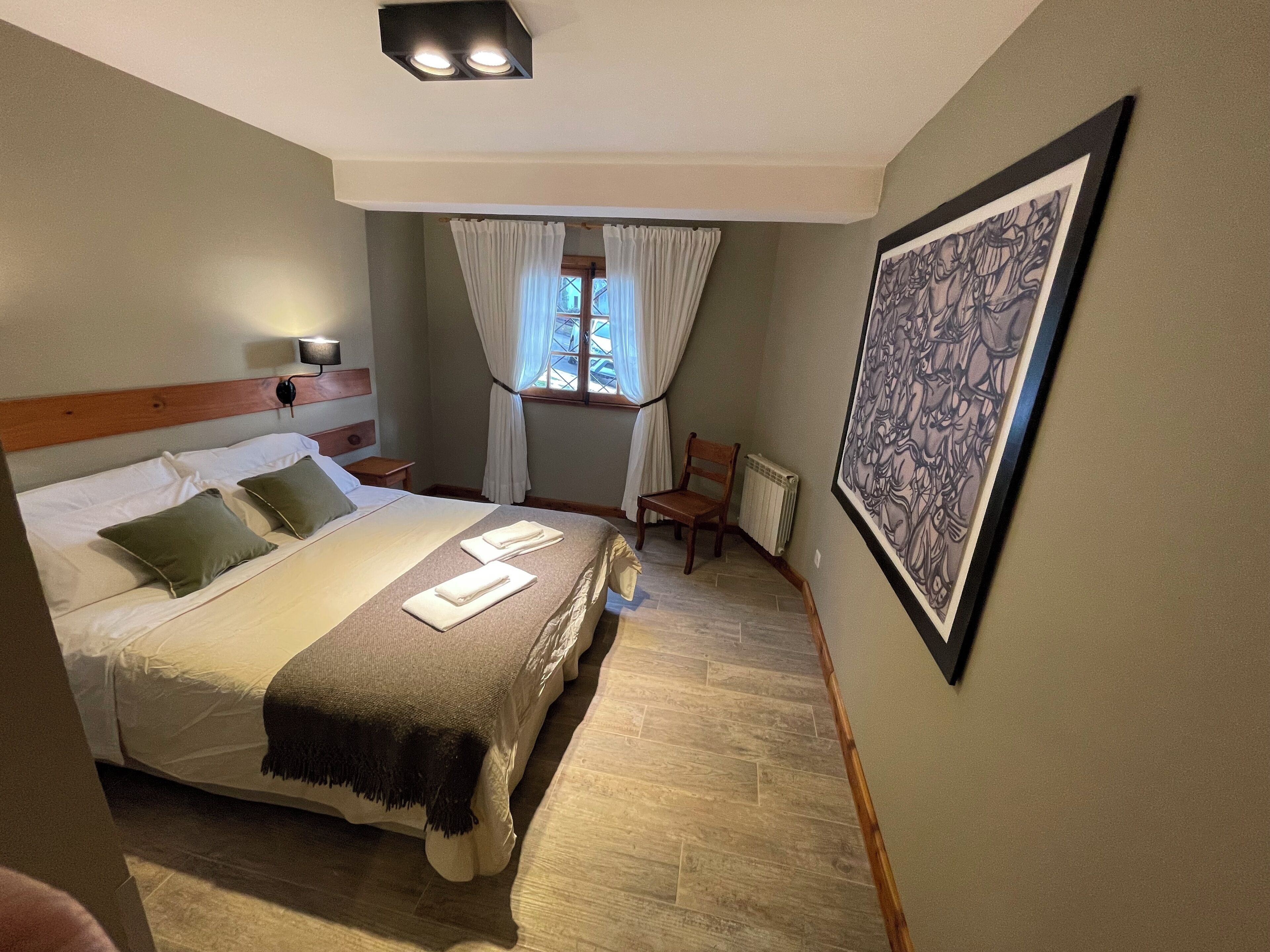A neatly made bed with pillows and a blanket, a bedside table with a lamp, a chair, a window with curtains, and a framed artwork on the wall.