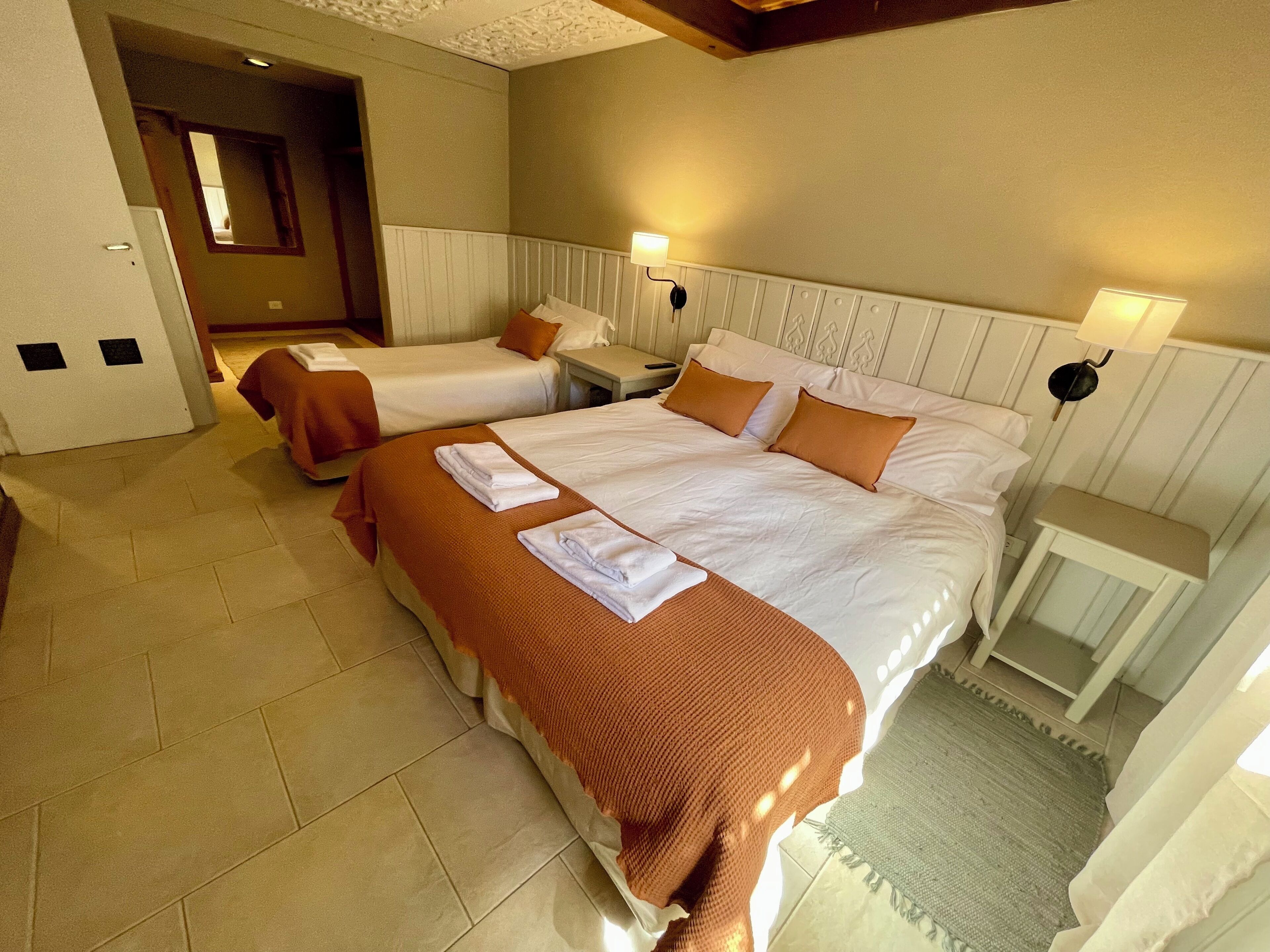 A hotel room with two beds, a bedside table, a nightstand, and a wall-mounted lamp.