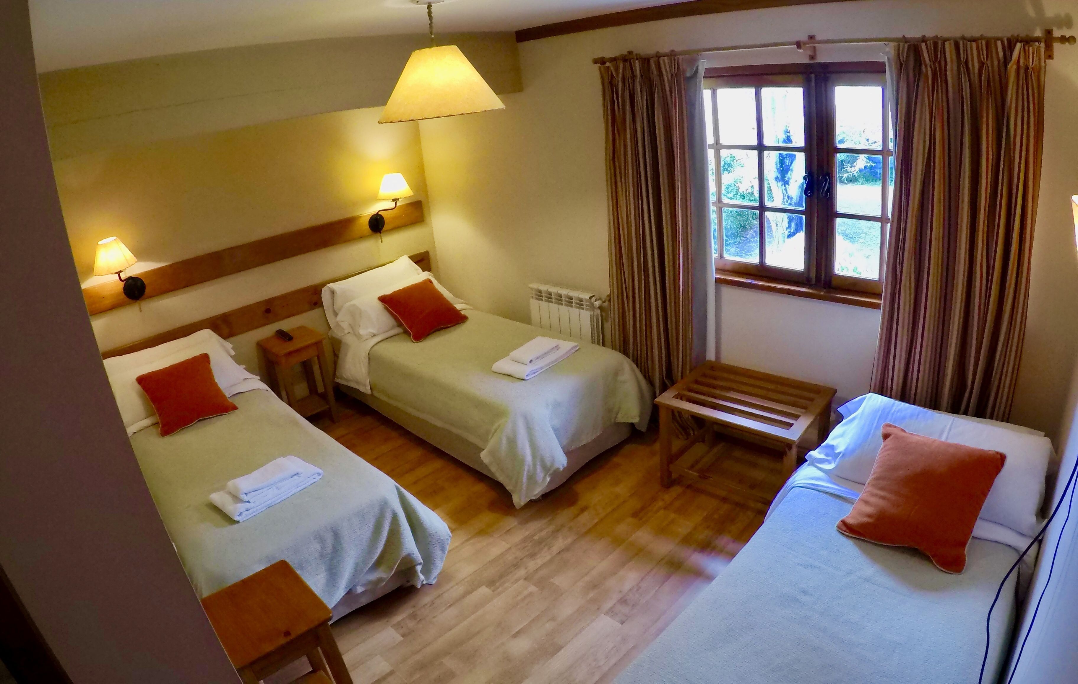 A hotel room with two single beds, a wooden bench, a table, a lamp, and a window with curtains.