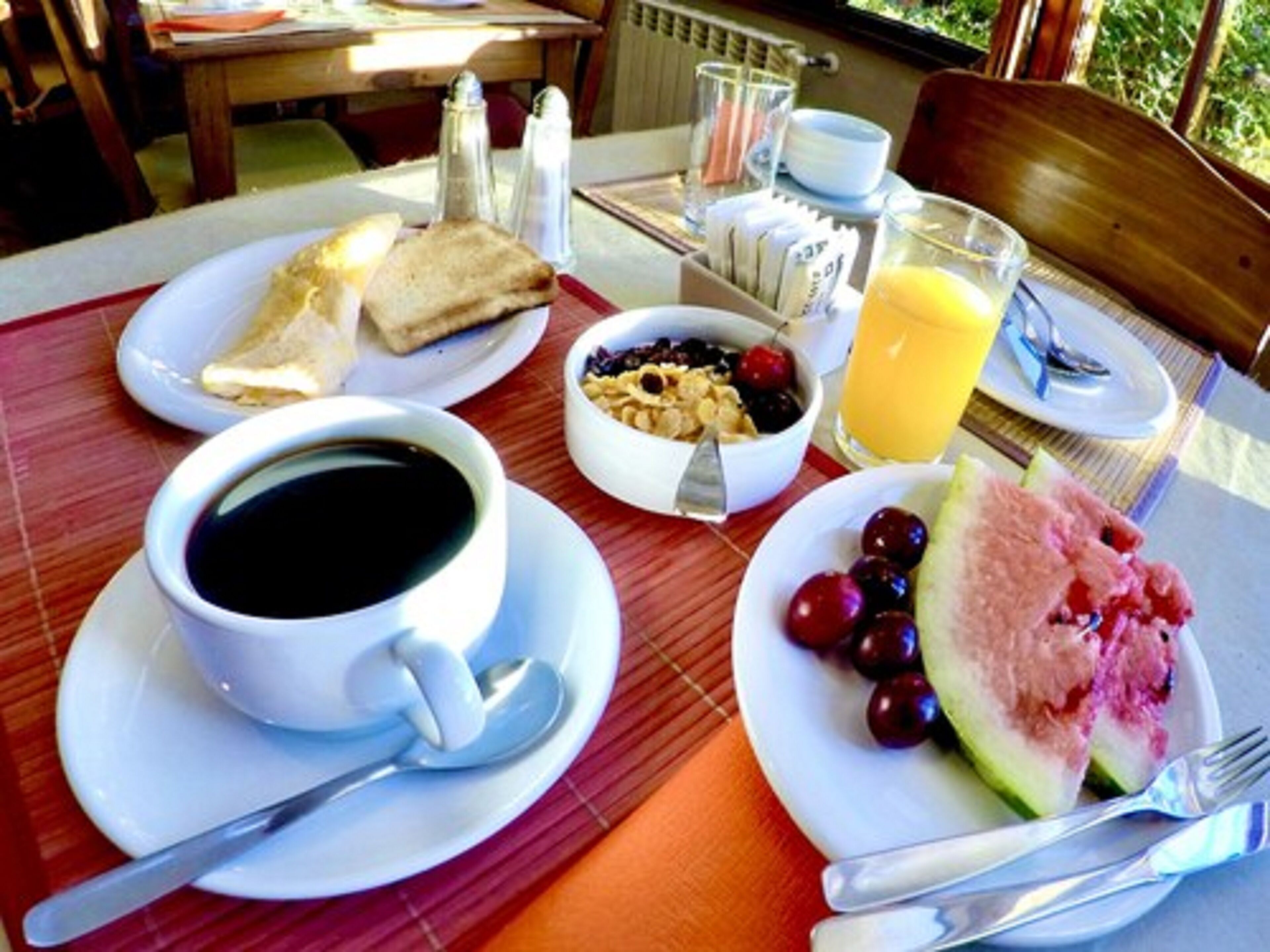 A breakfast table with a cup of coffee, a bowl of cereal, a glass of orange juice, a plate of fruit, and a slice of watermelon.