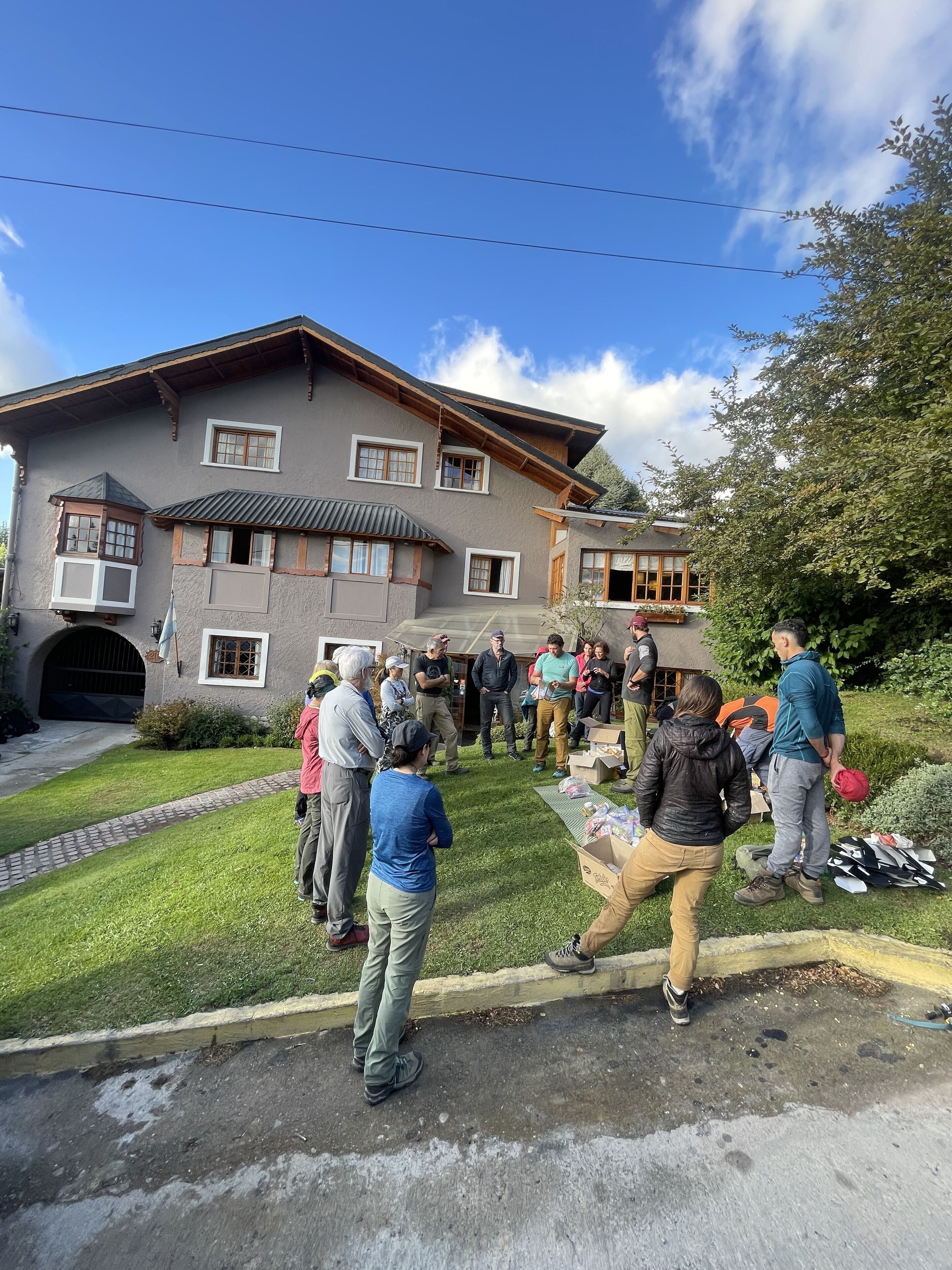 A group of people gathered outside a two-story house with a sloped roof and wooden beams, surrounded by greenery.