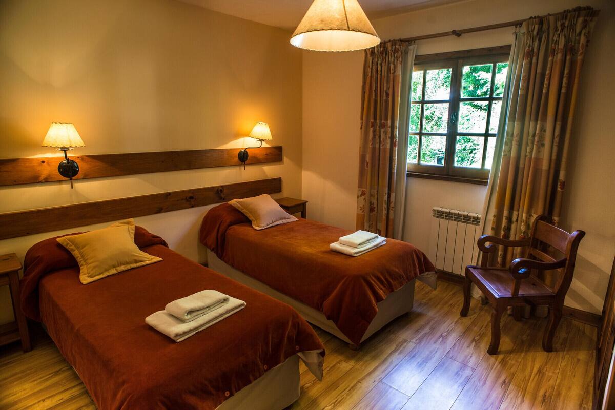 Habitación de hotel con dos camas, una silla, un ventanal con cortinas y lámparas fijadas a la pared.
