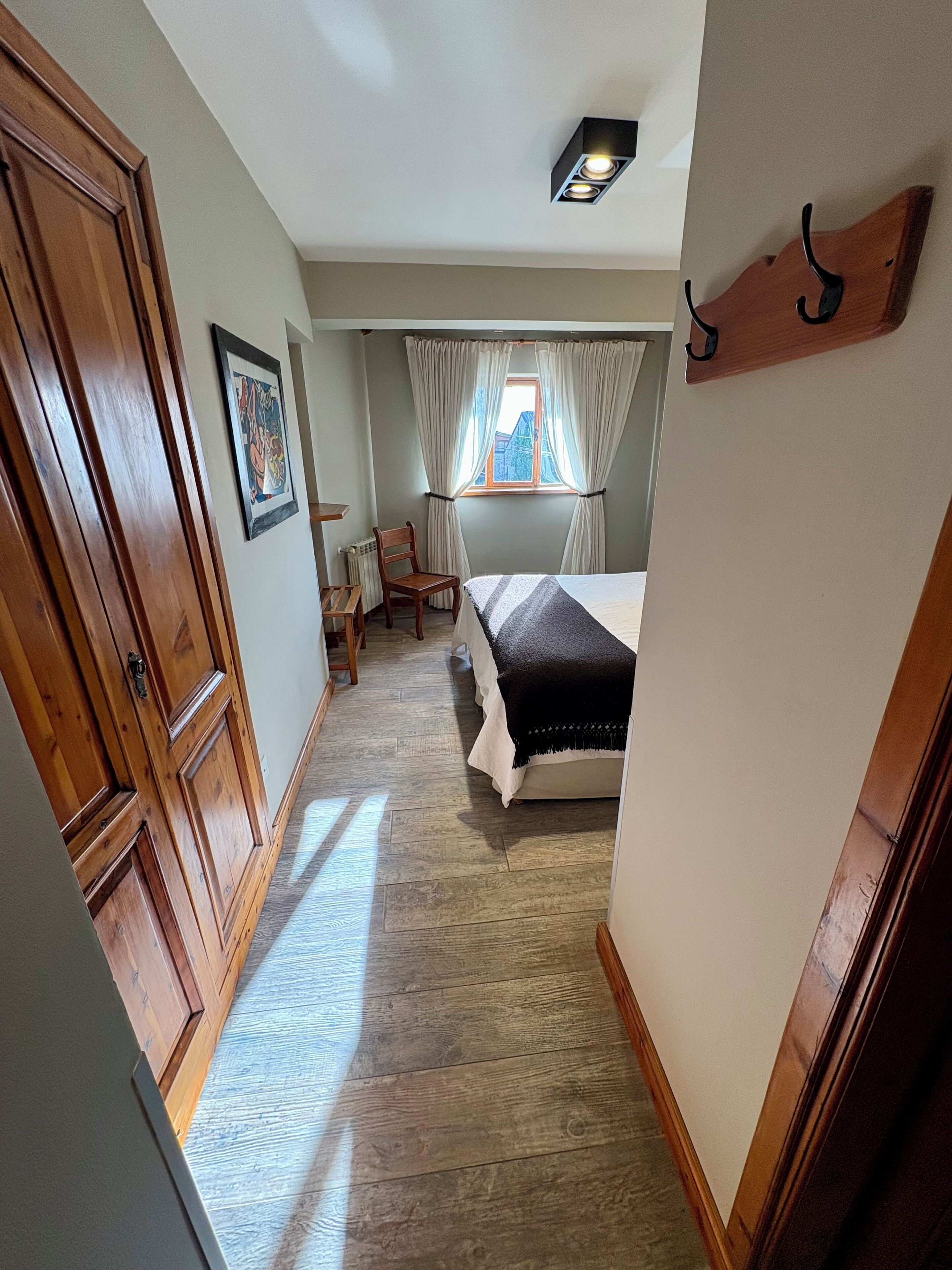 Un couloir étroit mène à une chambre qui contient un lit, une chaise, un radiateur, une fenêtre avec des rideaux et une porte en bois.