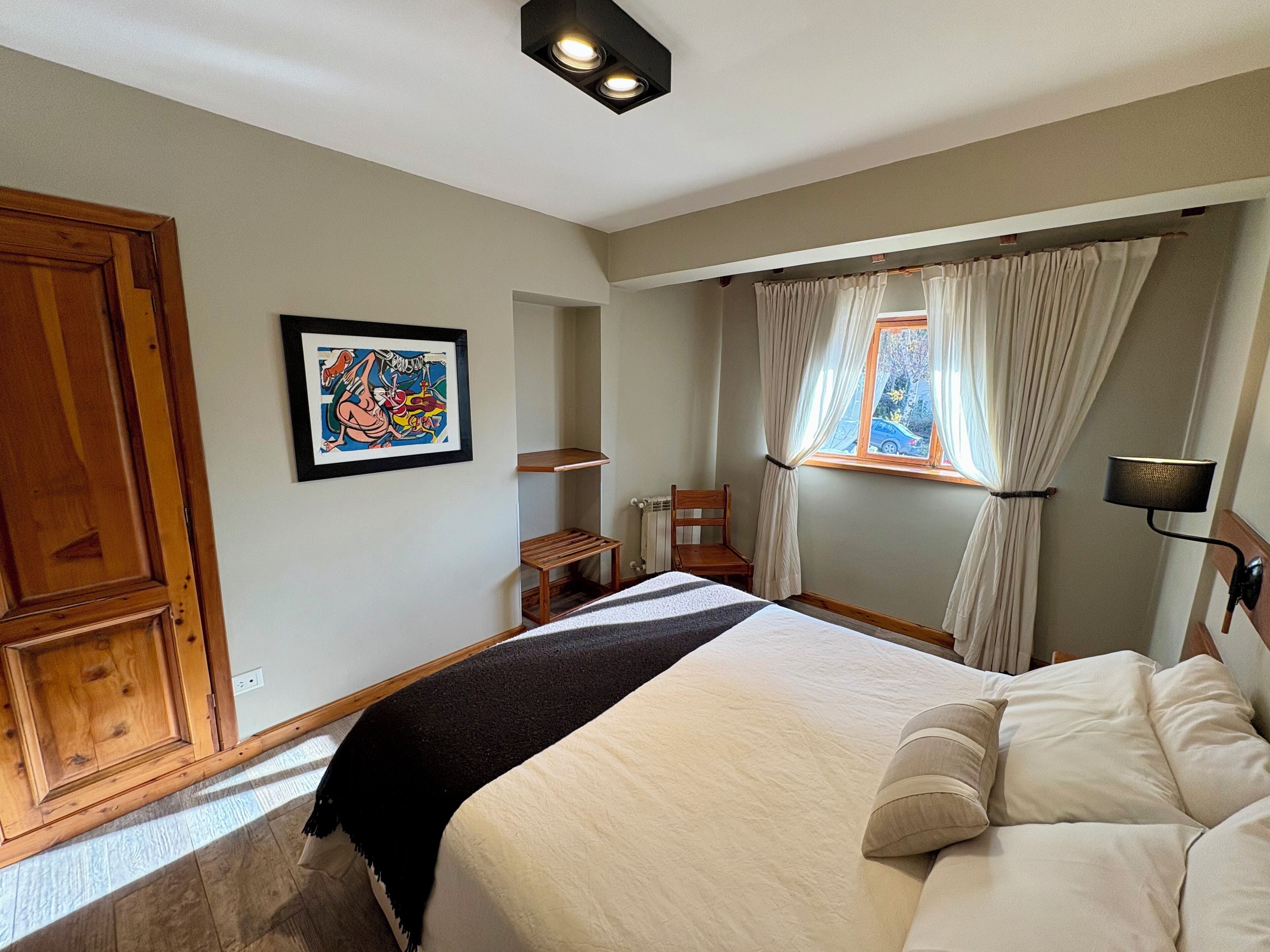 A bedroom with a bed, a chair, a small table, a window with curtains, and a wall-mounted lamp.