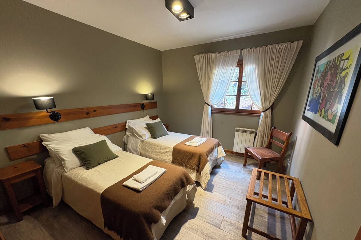Habitación de hotel con dos camas, cabecero de madera, mesita de noche, una silla, ventana con cortinas y un radiador.