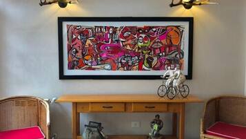 Una habitación con una consola de madera, sillas de mimbre con cojines rojos y un cuadro abstracto colorido sobre la mesa.