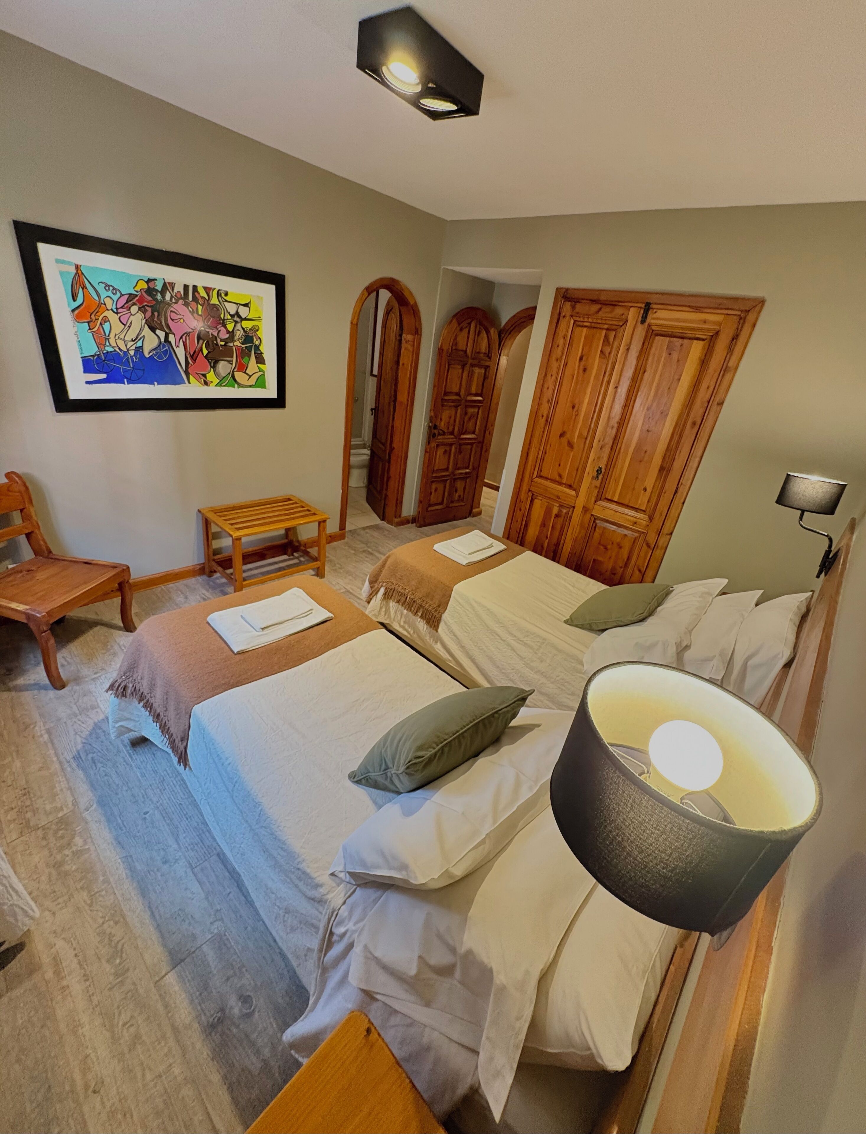 A bedroom with two beds, a wooden chair, a small table, a painting, and a lamp.