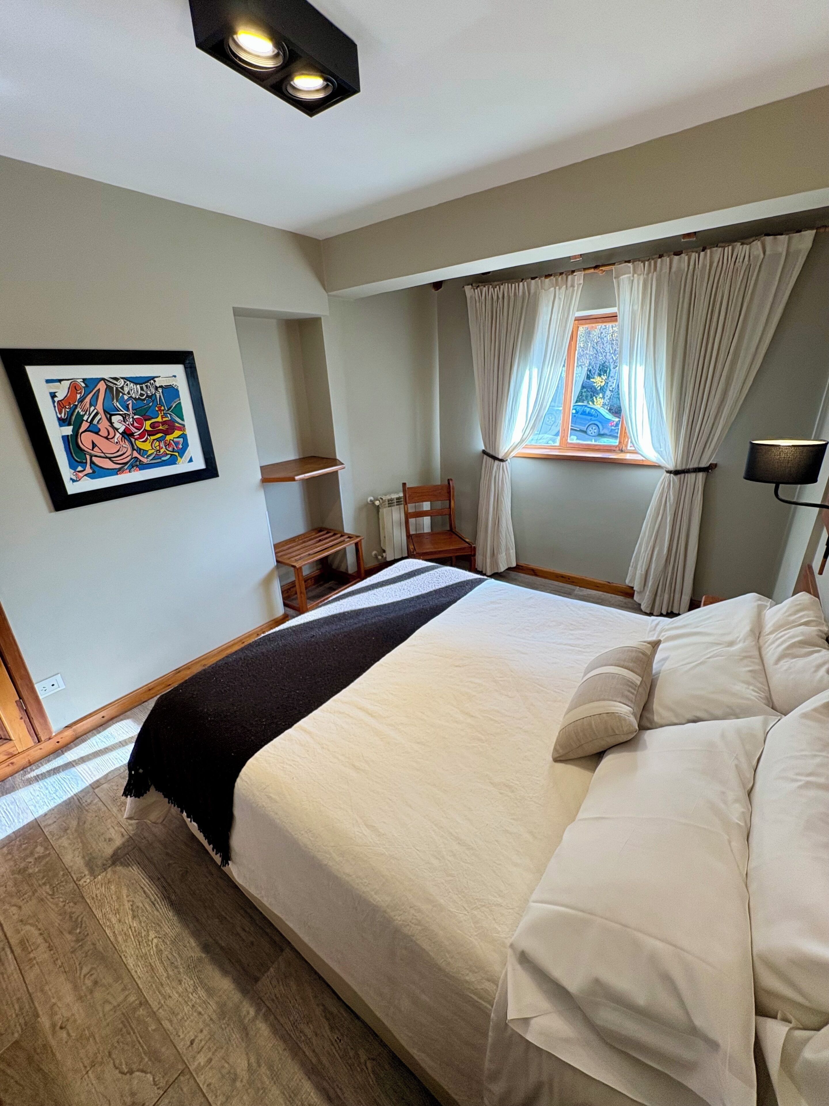 A bedroom with a large bed, a window with curtains, a radiator, a chair, and a framed picture on the wall.