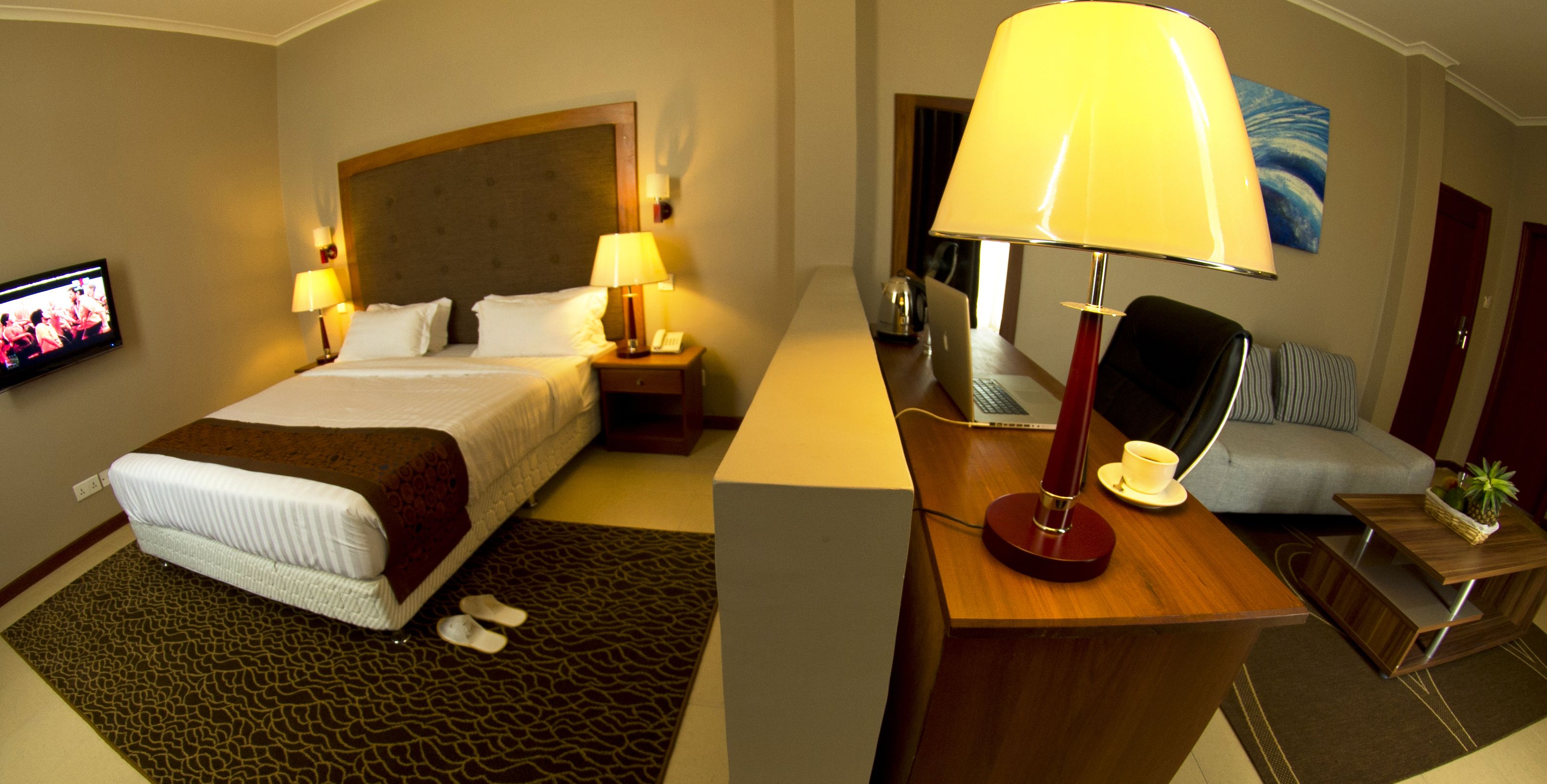 A hotel room with a bed, two bedside lamps, a desk with a lamp, a television, and a sofa.