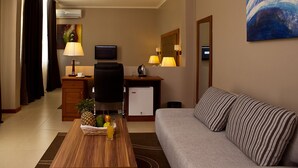 Una habitación de hotel con sofá, una mesa de centro con frutas y un vaso de jugo, escritorio con lámpara, televisión y un refrigerador pequeño.