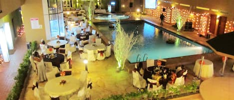 An outdoor dining area with tables and chairs, a pool, and a building with lights in the background.